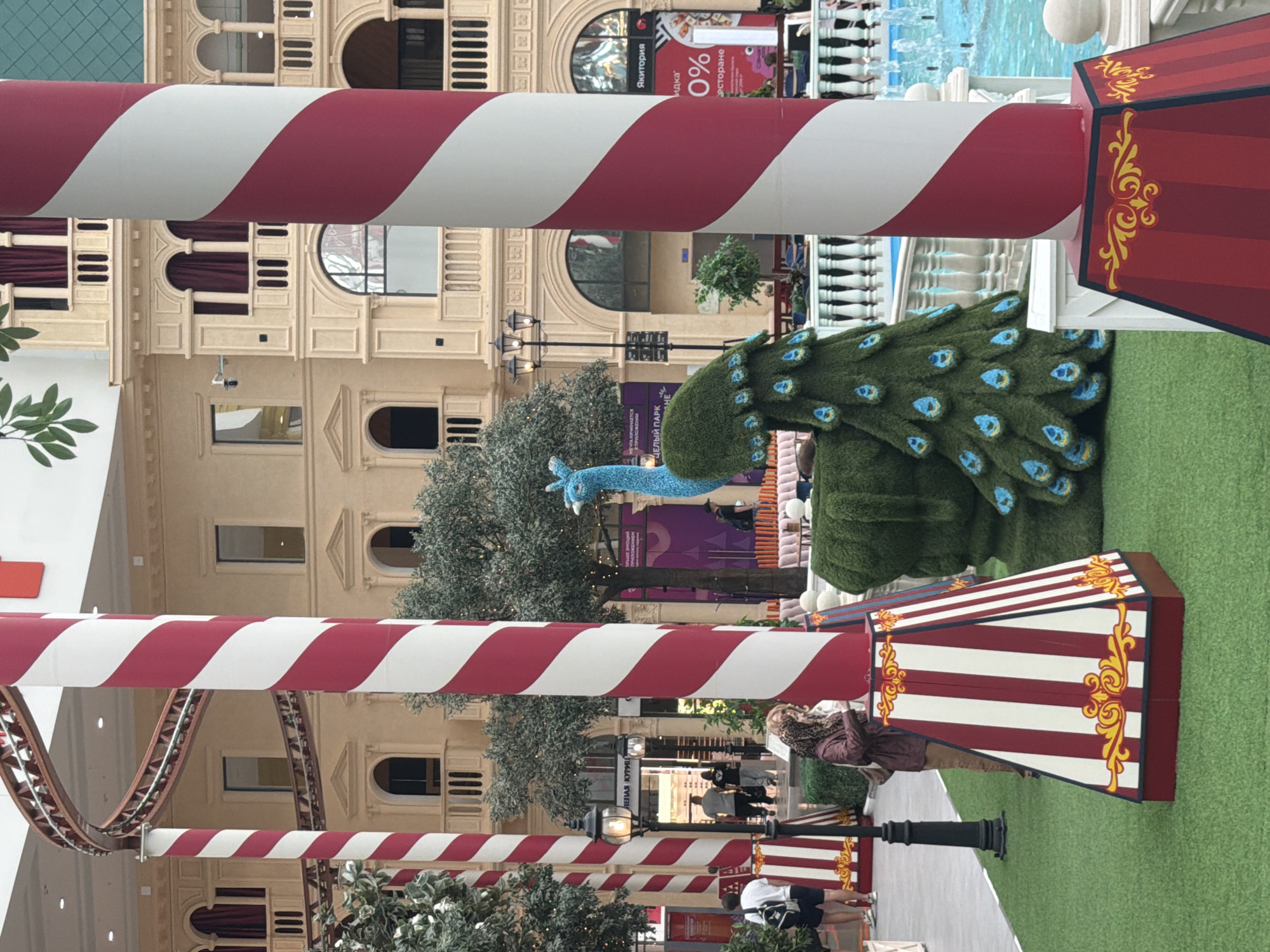 The image depicts an indoor scene, possibly a mall or entertainment venue, with a strong emphasis on decorative elements. Large, red and white striped poles dominate the foreground, creating a striped pattern that runs through the frame. To the right, a decorative, tiered structure with red and white stripes and gold ornate designs is visible, resembling a circus-themed booth or stand.
In the midground, a sculpted peacock is prominently displayed. It is made of green material, designed to mimic foliage, and features blue and black "eyes" on its tail feathers. A bright blue peacock head and neck extends from the body. Behind the peacock, a grey, leafy artificial tree stands.
The background reveals architectural details of a building with a light-colored facade, featuring arched windows and decorative elements. Signs are visible in the distance, one with red lettering against a white background that reads "ЯКИТОРИЯ" and another with "0% РАСТОРАНЕ". There are also glimpses of people walking in the background, suggesting the venue is open to the public. One person in the mid-right is wearing a brown jacket and a leopard-print scarf, and another person in the foreground is wearing shorts and a white t-shirt. The overall impression is of a festive or decorative display, possibly for an event or holiday. The lighting suggests it is daytime, and the indoor setting implies controlled weather.