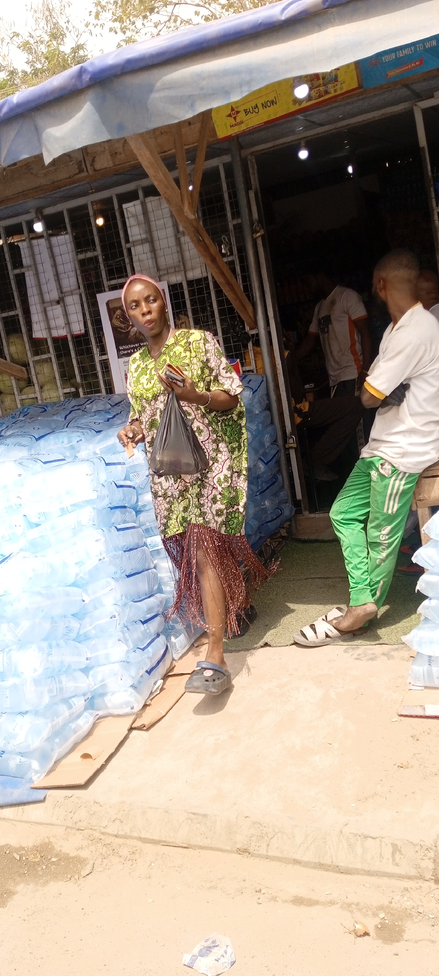 Outside a shop in Gayawa, Nigeria, a woman with a pink head wrap wearing a green and white patterned dress, brown fringed hem, and black Crocs stands next to a stack of bottled water. She is carrying a black plastic bag and a wallet or phone. To her right, a man in a white shirt and green Adidas pants leans against the shop’s entrance. Inside the shop, other people are visible. Signs are displayed, including a yellow Maggi advertisement and a blue sign with the phrase "YOUR FAMILY TO WIN." The ground is sandy and there's a piece of trash.