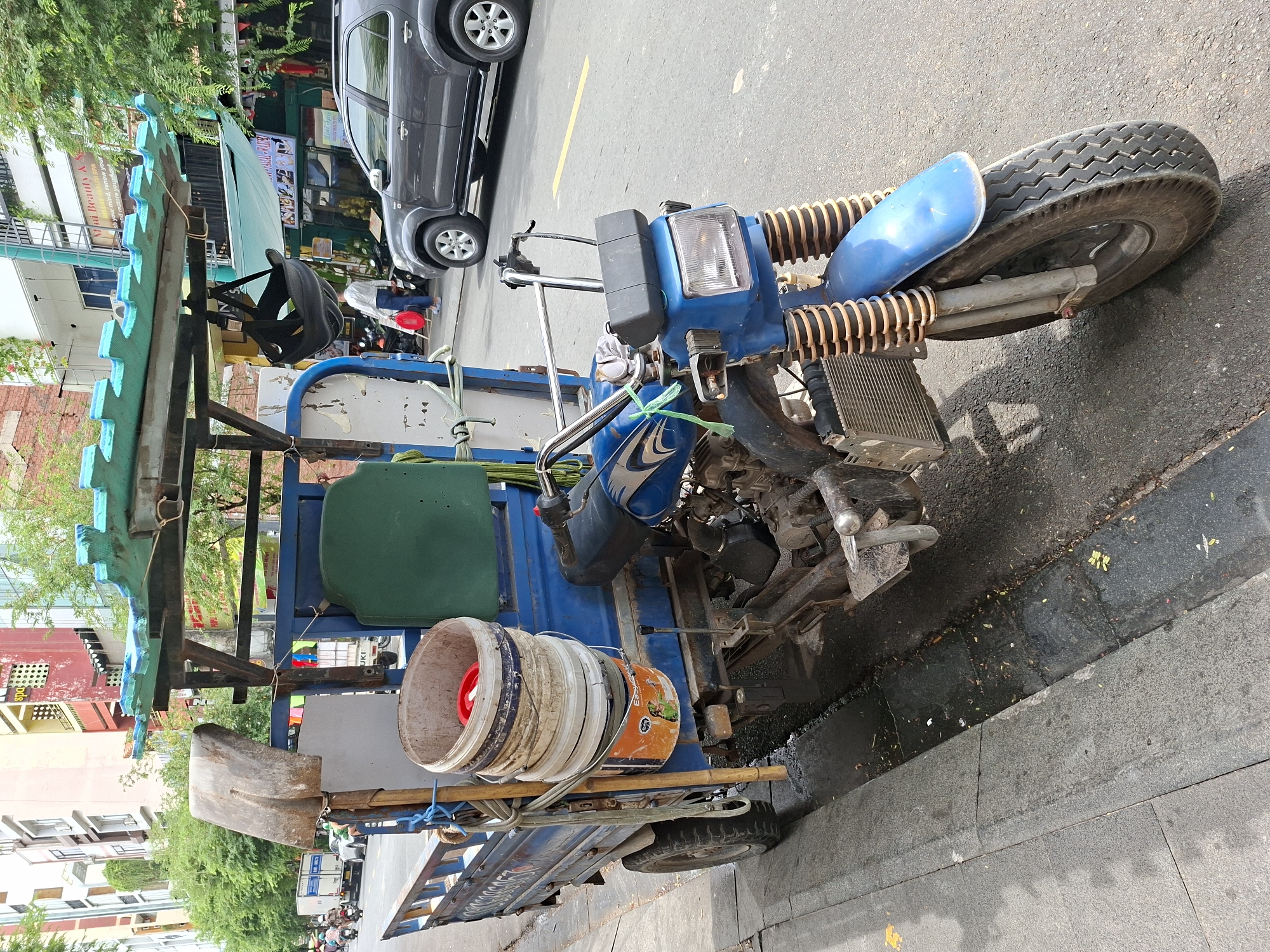This is a medium shot, taken from a slightly low angle, of a three-wheeled motorized cart parked on the side of a street in Thủ Đức City, Vietnam. The cart is blue and has a light green, corrugated plastic roof supported by metal poles. Attached to the front of the cart is a blue motorcycle with a black seat. The motorcycle has chrome handlebars and a black headlight assembly. The front of the cart has a metal frame extending forward, with what appears to be a radiator or cooling fins.
On the rear of the cart, there is a stack of five white and orange buckets, with a red lid on the top bucket. To the left of the buckets, a shovel with a worn metal blade is propped upright. A bamboo pole is also tied to the side of the cart, along with various ropes and straps. A green plastic seat is visible inside the cargo area of the cart. A black helmet is hanging from the roof structure.
The street appears to be paved, with a sidewalk made of dark grey stone tiles running along the left side of the image. The street is wet in places, with some debris and fallen leaves visible on the asphalt. In the background, there are buildings with colorful facades, trees, and other vehicles, including a dark gray SUV parked on the street and several motorcycles further down the road. One person, wearing a white shirt and dark pants, is walking away from the cart carrying a red bag. The scene appears to be typical of an urban street in Vietnam, likely during the daytime given the lighting.