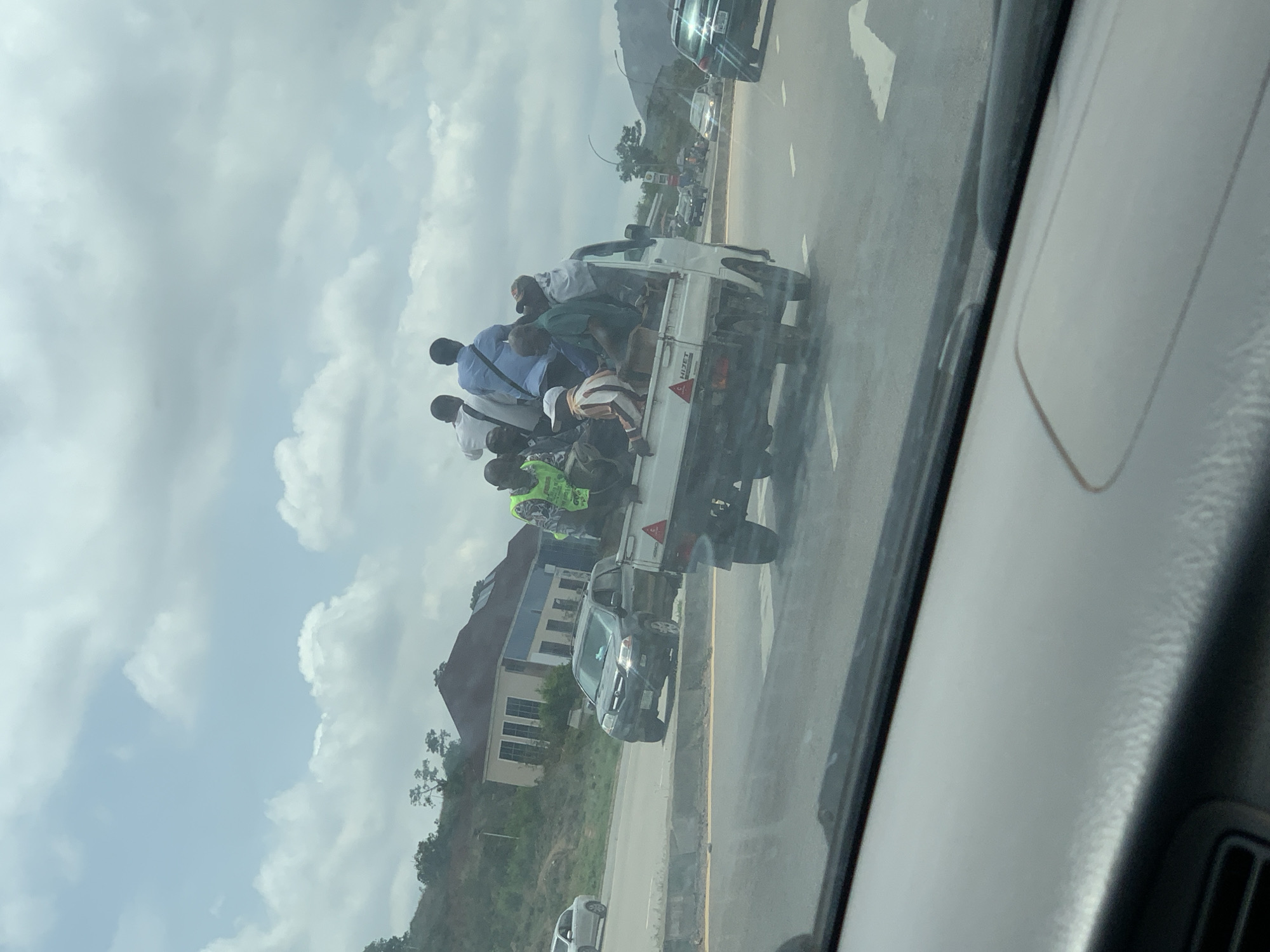 A photograph taken from inside a moving vehicle shows a white pickup truck on a highway in Nyanya, Nigeria. The truck bed is filled with approximately 15 people, some standing and some sitting. They appear to be men, dressed in a mix of casual and business attire. A man near the rear of the truck is wearing a high-visibility vest. Other vehicles are on the road in the background, along with buildings and green, hilly terrain. The sky is cloudy.
