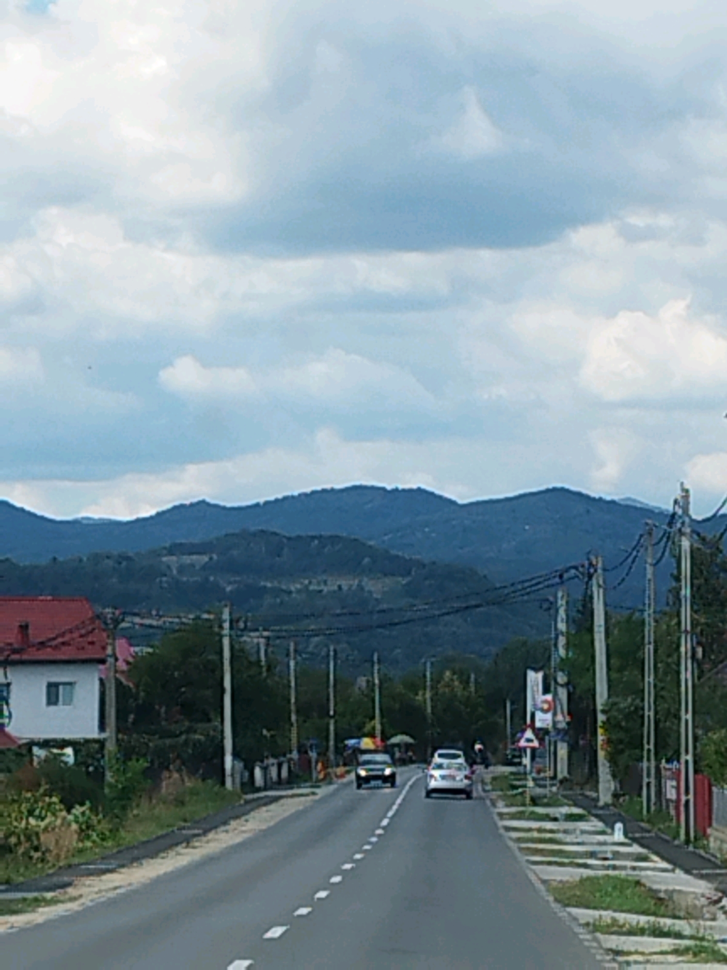 The image depicts a straight, paved road in Albeștii Ungureni, Romania, on an overcast day. The road is lined with dashed white markings. On the left side of the road, a white house with a red roof is visible, situated behind a patch of green vegetation. Further down the road, two cars are traveling in the same direction: a dark-colored car followed by a white car. On the right side of the road, there are several utility poles with numerous wires strung between them, along with some trees and a red fence. In the background, a range of dark, undulating hills or mountains under a cloudy sky dominates the horizon. The overall scene suggests a rural or semi-urban setting.