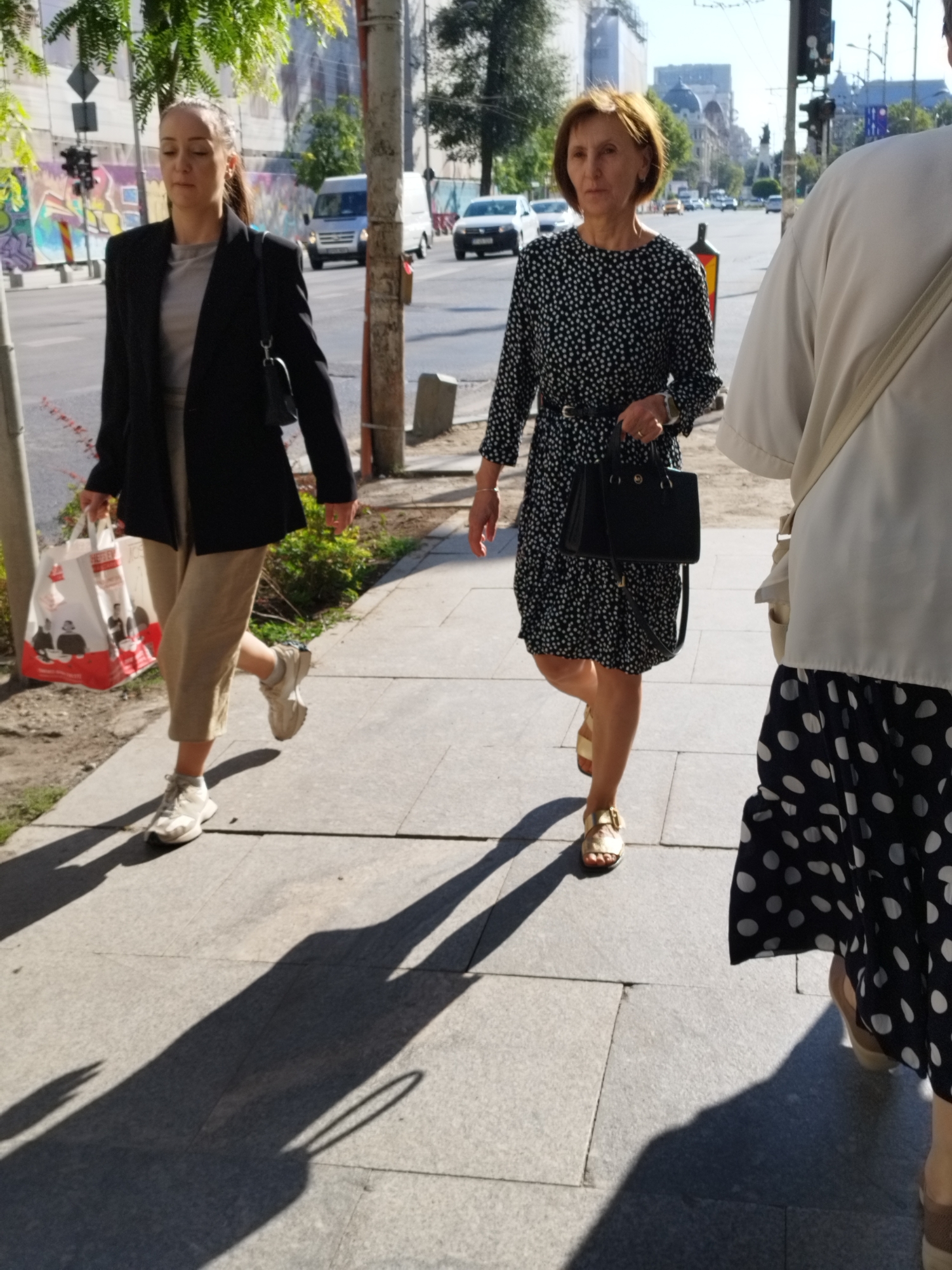 This image captures a street scene in Bucharest, Romania, with three individuals walking on a sidewalk. The foreground features the lower body of a person on the right, wearing a white top and a black skirt with white polka dots, partially obscured by shadow. To their left, a woman in a black and white polka dot dress with a black belt walks towards the viewer, carrying a black handbag. Her right foot is visible, wearing a gold sandal. Further left, another woman, wearing a black blazer over a grey top and beige cropped pants, walks away from the viewer. She carries a large white shopping bag with red accents. The sidewalk is made of grey paving stones, with shadows cast by the pedestrians and trees. The background shows a street with traffic, buildings, and foliage, suggesting an urban environment.