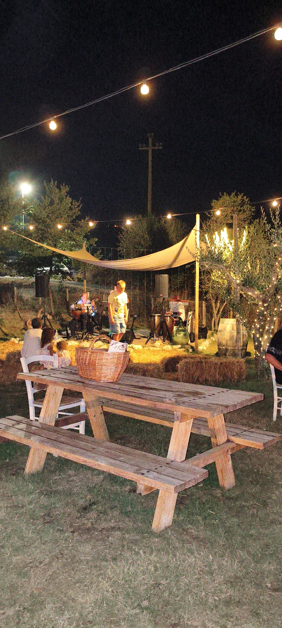 An outdoor evening scene is depicted in Buriano, Italy, featuring a rustic wooden picnic table in the foreground with a woven basket on its surface. Behind the table, a live band performs on a raised platform covered by a light-colored canopy. Several individuals are present, with some seated in the audience and others on stage. String lights are strung overhead, illuminating the area, and trees adorned with more lights flank the performance space. Hay bales are visible behind the band, and a wooden barrel stands to the right. The overall ambiance suggests a casual outdoor gathering or concert.