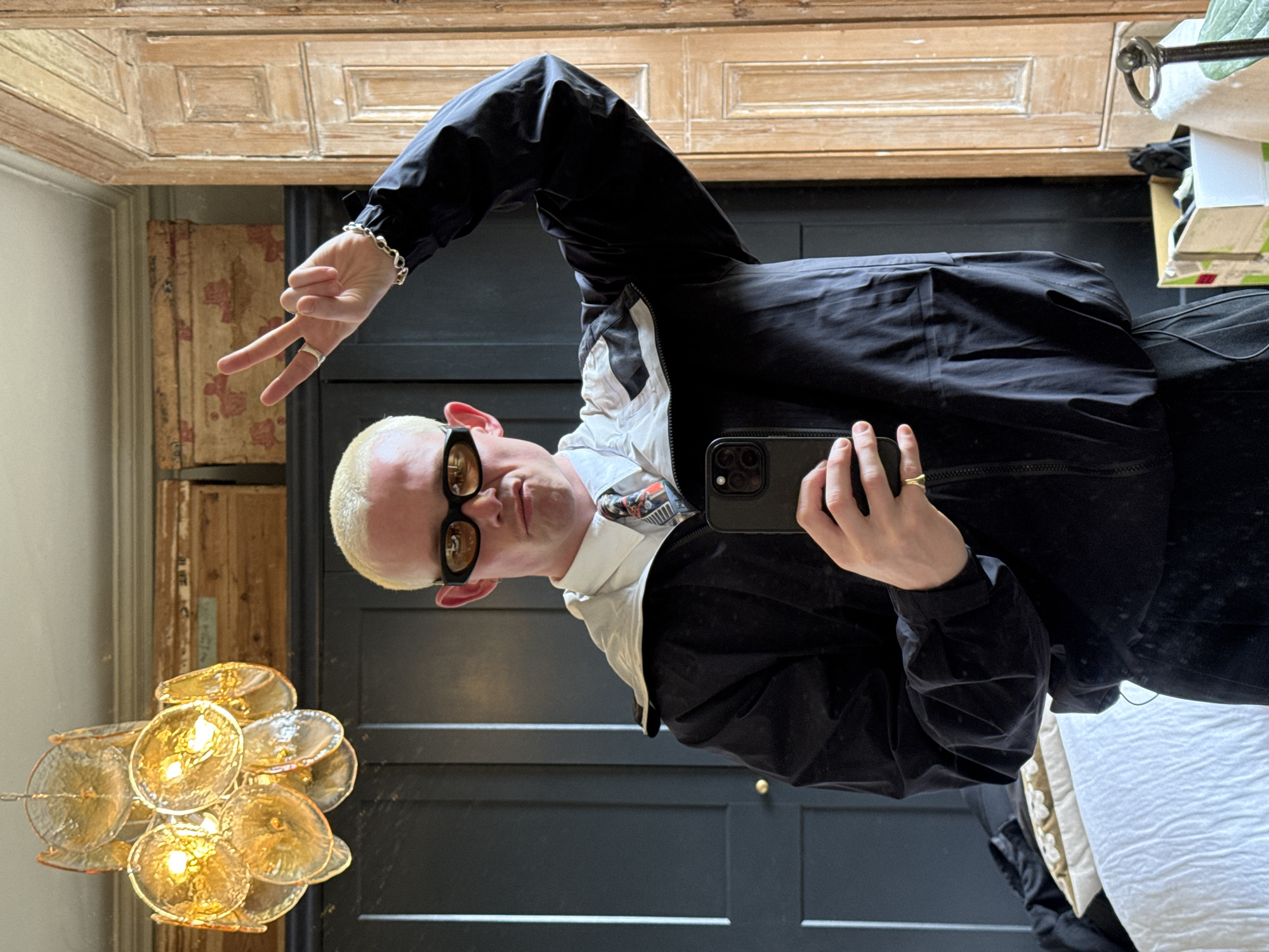 A medium shot depicts a person indoors. The person is fair-skinned with blonde hair and is wearing sunglasses, a black jacket, and a white collared shirt. They are holding a phone in their right hand, seemingly taking a selfie, and extending their left hand, making a peace sign. The background includes a dark paneled door, a wooden cabinet above, a chandelier hanging on the left, and a glimpse of bedding on the right. The location cannot be determined based on this image alone, but it is specified that this image is from London, United Kingdom.