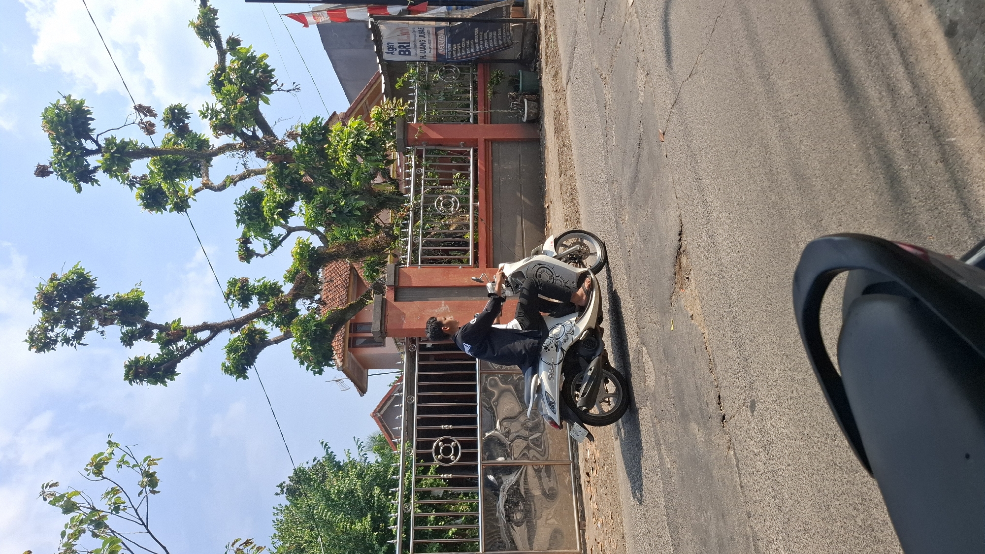 A young man rides a white scooter on a paved road in Cilaku, Indonesia. He is wearing a black long-sleeved shirt and dark pants, with his right foot on a pedal and his left foot on the ground. The scooter is decorated with a pattern of swirling designs. In the background, a metal gate with ornate circular designs separates a paved area from a street. Behind the gate, a red-tiled roof house is visible, with large trees with sparse foliage casting shadows on the ground. To the right of the gate, a sign for "Agen BRI" and "K. UANG JUMLAH" is mounted vertically on a pole. The sky is clear blue with scattered white clouds. The road surface is weathered and cracked, with some potholes. In the foreground, the rear of another black scooter is visible.