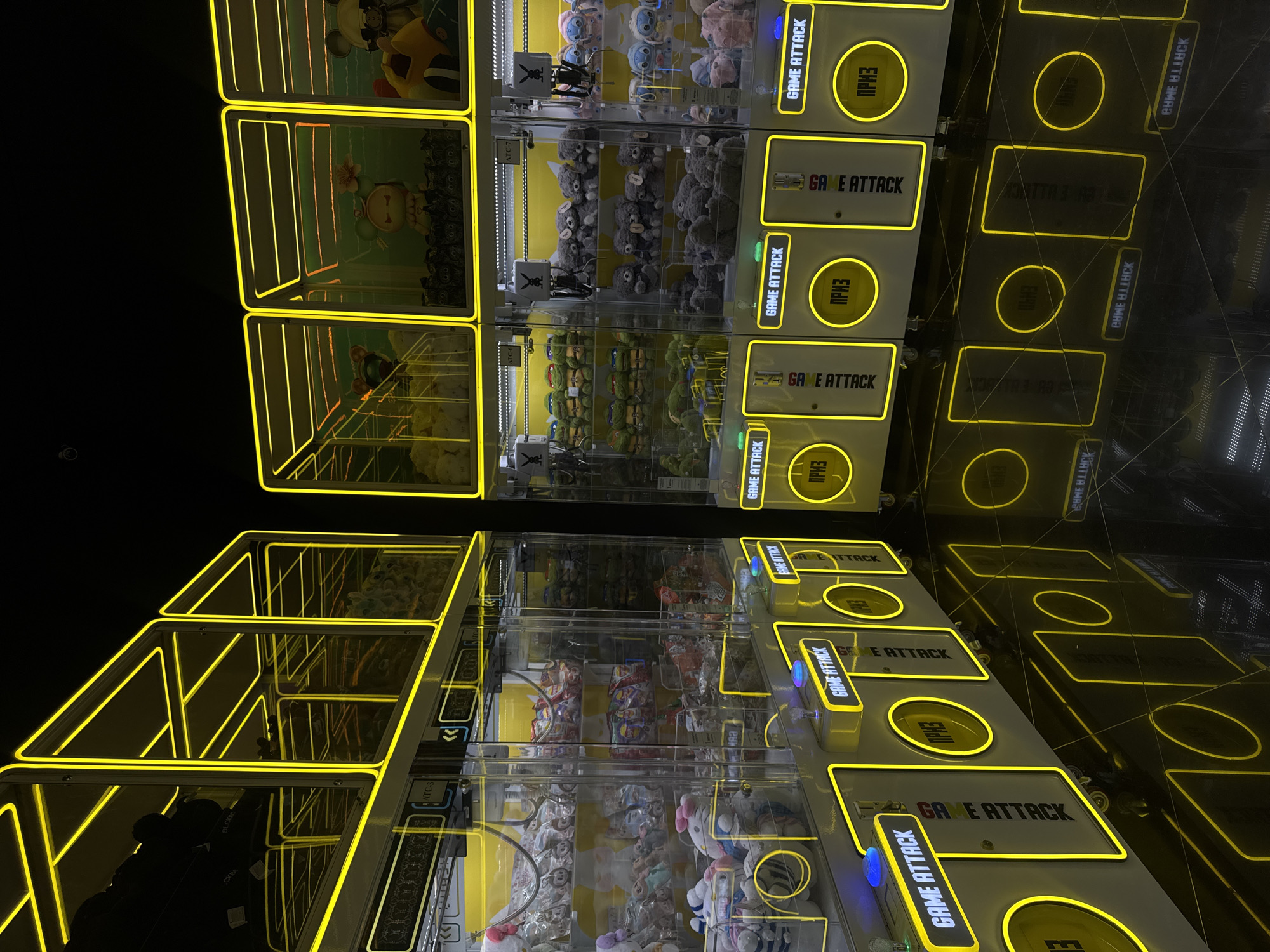 The image shows a set of prize claw machines in what appears to be an arcade or entertainment venue. The machines are brightly illuminated with yellow neon-like lights outlining their structure. The machines' interiors contain various plush toys and prizes, including licensed characters like ninja turtles. The reflection on the floor suggests a polished surface. The language on the machines is a mix of English ("GAME ATTACK") and what appears to be Russian Cyrillic. Considering the Cyrillic writing and assuming it is a commercial arcade, this photo could plausibly be taken in Moscow, Russia. No people are clearly visible, although a dark form appears in the bottom left, potentially a person. The environment is dark, with the bright lights of the machines providing the main source of illumination.
