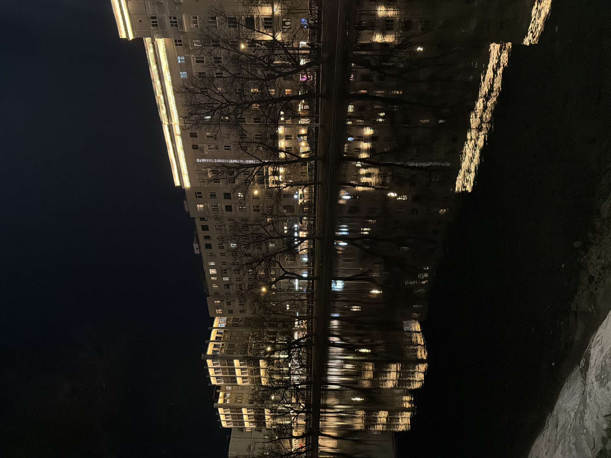 This image presents a tranquil night scene of an urban environment, identified as Moscow, Russia. The central focus is a long, multi-story building illuminated by artificial lights, stretching horizontally across the frame. The building features bright, warm-toned strip lighting along its roofline and prominent horizontal architectural elements, alongside numerous glowing windows that suggest activity within.
In front of the building, several bare deciduous trees, characteristic of winter, stand silhouetted against the bright facade, their intricate branch structures visible. Below the building and trees, a dark body of water – likely a canal or river – creates a prominent mirror effect, reflecting the building's lights and the shadowy forms of the trees. These reflections are slightly distorted by gentle ripples on the water's surface, adding a dynamic texture to the otherwise still scene.
The sky above is a deep, unlit expanse of night, serving as a dark backdrop that intensifies the luminosity of the building. To the right, a darker, textured bank or wall borders the water. The scene is quiet and still, emphasizing the beauty of urban lighting reflected in water. There is no visible text in the image.
