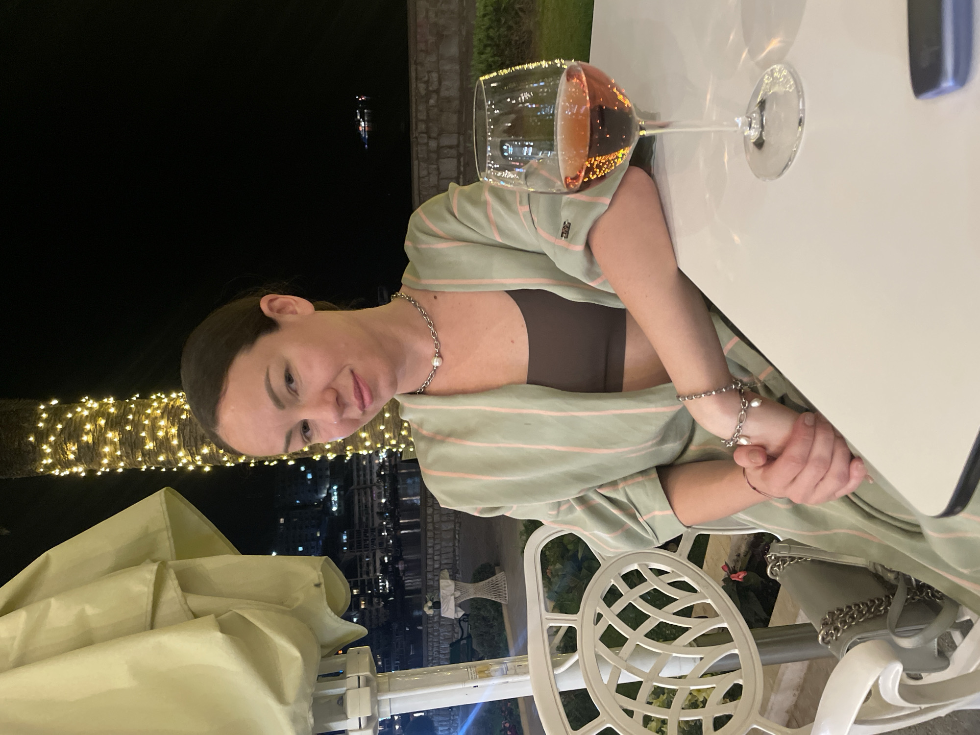 A woman sits at a table outdoors at night in Rafailovići, Montenegro. She wears a pale green, striped robe over a brown top, and has a silver necklace and bracelet. Her hands are clasped on the white tabletop next to a glass of rosé wine. In the background, lights decorate a palm tree, and the dark outlines of buildings and the coastline are visible. A white, lattice-style chair and a light-colored patio umbrella are also visible. A small gray purse with silver chain is on the chair.