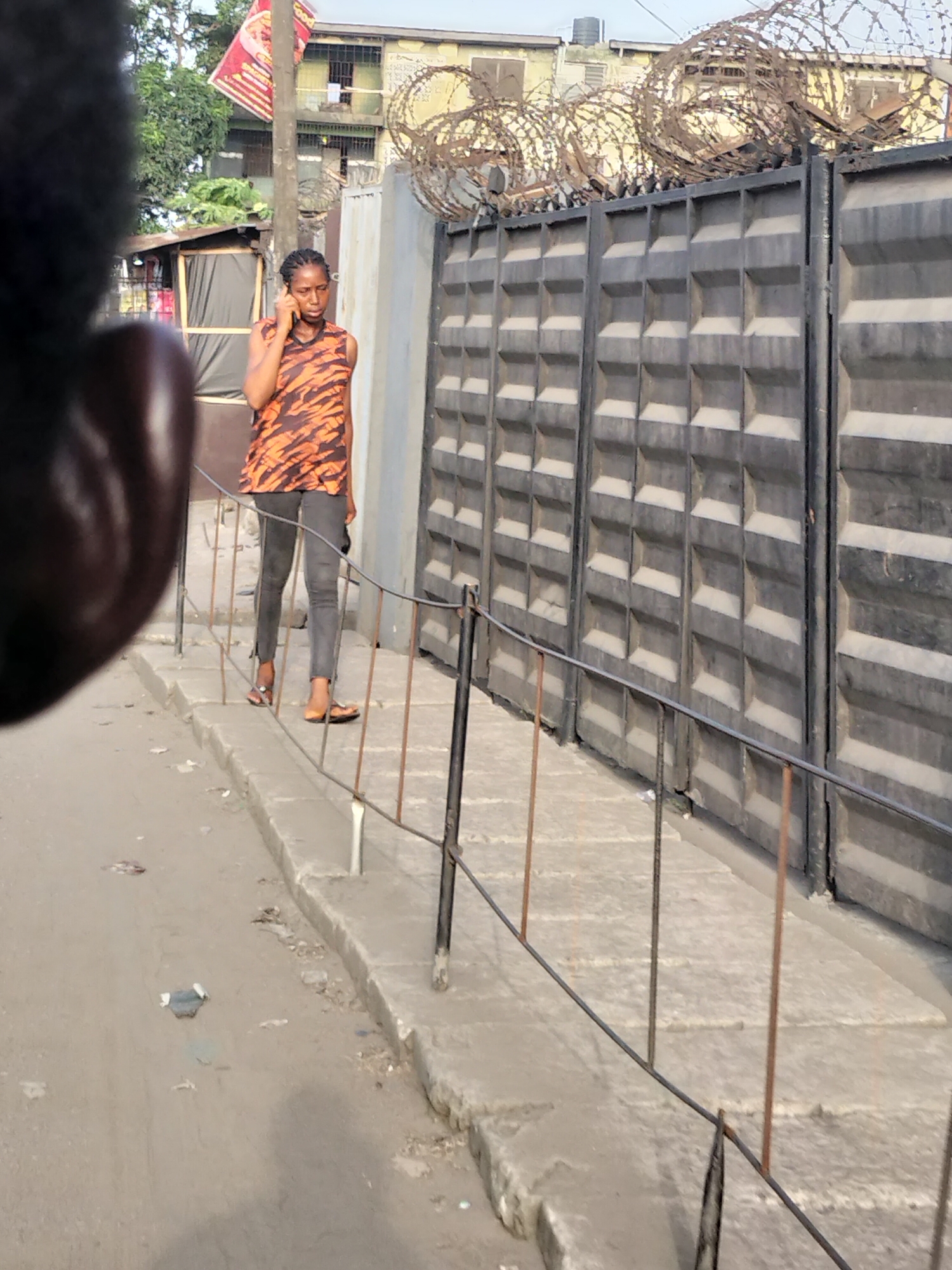 The image shows a street scene in Alaiyabiagba, Nigeria. A woman is walking along a concrete sidewalk while talking on a cell phone. She is wearing a brown and orange animal print sleeveless top, gray pants, and brown sandals. A large gray gate with barbed wire on top runs along the sidewalk. The gate appears to protect a multi-story building in the background. A metal fence runs along the edge of the sidewalk. The street appears to be paved and has some trash and debris scattered on it. A blurred dark object is in the extreme left of the frame. A sign is visible in the background.