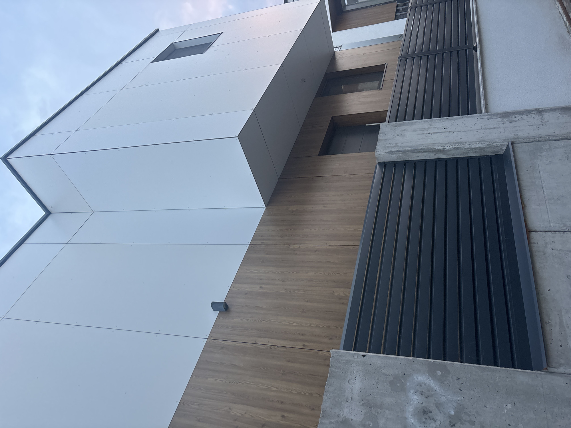 The image depicts the exterior of a modern building, likely a residential property, in Burgas, Bulgaria. The building features a two-toned facade: the upper section is clad in large, white panels with visible seams, and the lower section is covered in vertical wooden panels with a prominent grain pattern. A dark, horizontal slatted fence with concrete posts runs across the foreground, separating the building from a white-painted wall. A small, dark-colored window is visible on the upper right section of the building, and a darker doorway is set within the wooden-clad section. The sky above is overcast and appears to be twilight or a cloudy day. There are no people or animals visible in the frame.