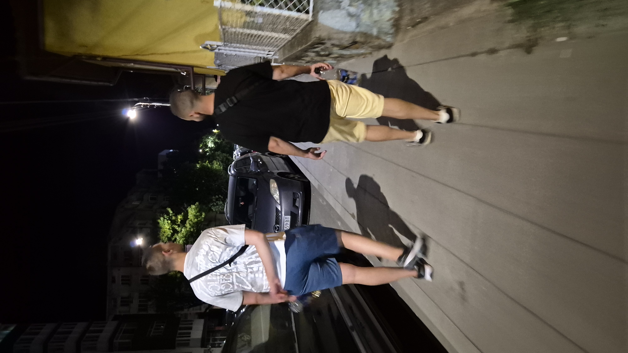 Two men are walking away from the camera on a sidewalk at night. The man on the left is wearing a white t-shirt with graphics and blue denim shorts. The man on the right is wearing a black t-shirt and yellow shorts. Both are wearing athletic shoes. They are walking on a paved sidewalk next to a street with parked cars. In the background, there are apartment buildings and trees, illuminated by streetlights. The scene appears to be in a residential area. The location is Lovech, Bulgaria.