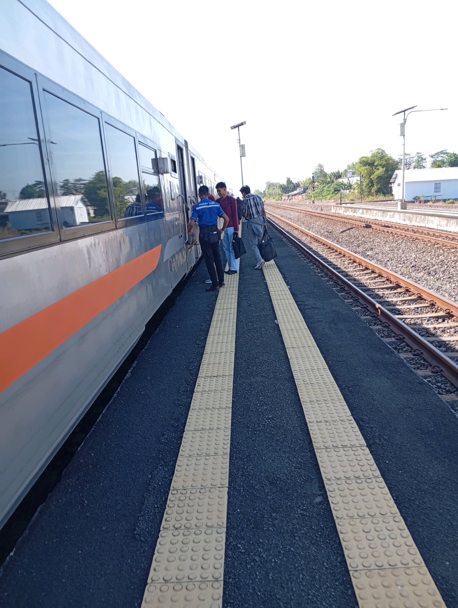 At a train station in Tebon, Indonesia, a silver train with an orange stripe is stopped. Three individuals stand on the platform. One person, wearing a blue shirt with "FACILITY CARE" printed on the back, appears to be a staff member and is facing the train. Another individual in a striped shirt and gray pants is walking away from the train, carrying a black bag. A third person, in a maroon shirt, stands behind the individual in the blue shirt, looking towards the train. The platform has a textured yellow safety strip running along its edge. Train tracks are visible next to the platform, and a small white building is in the background. The sky is clear and bright.
