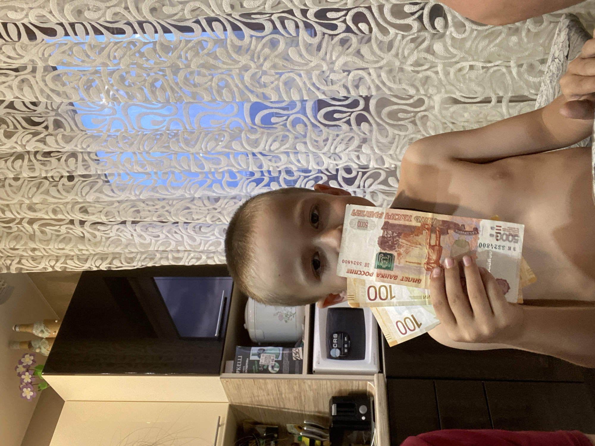 A young boy is holding Russian banknotes in front of his face. He has short, light-colored hair, and his torso is visible. He is standing in front of a window covered by a white patterned curtain. To the left of the boy is a black cabinet with drawers containing various items. The banknotes in his hand include at least one 5000 ruble note and two 100 ruble notes. The environment appears to be indoors, possibly a home in Марковский, Russia.