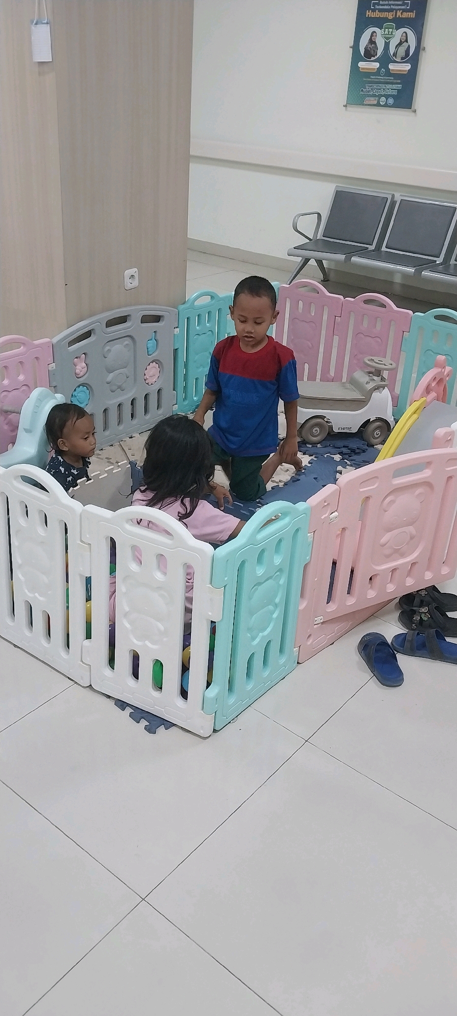 In the city of Kebonagung, Indonesia, within a building resembling a waiting area, three young children are playing in a colorful plastic playpen. The playpen is comprised of interlocking panels in white, pale pink, and light blue, some decorated with embossed animal shapes. Inside the playpen, a young boy wearing a blue and red striped t-shirt and green shorts kneels, appearing to interact with a young girl wearing a pink shirt. Another very young child, partially visible, sits to the left, also within the playpen. The playpen floor is covered with interlocking foam mats. Scattered outside the playpen are several pairs of sandals. In the background, a row of grey chairs suggests a waiting area, and a poster with text and images is mounted on the wall. The environment appears to be an indoor public space.