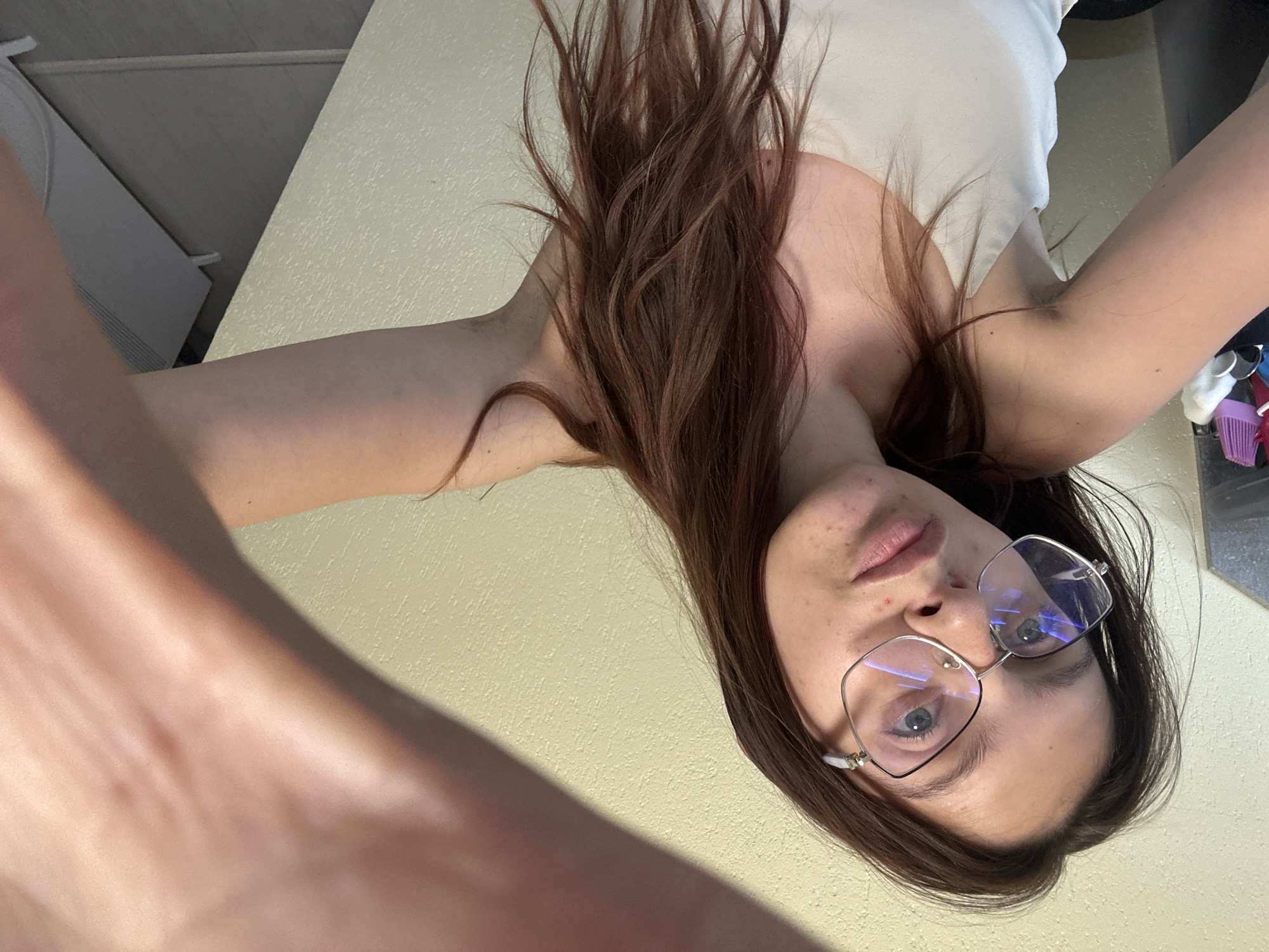 A young woman with long, dark brown hair and glasses is taking a selfie. She is wearing a white tank top. Her right arm is extended towards the camera, partially obscuring the scene. The background appears to be a room with a textured, light yellow wall and a grey floor. In the background, a white heater or air conditioner unit is visible. The woman's face is turned slightly to the left, and her gaze is directed towards the camera. There are no specific landmarks or features that would allow for the identification of the city of Tryavna, Bulgaria.