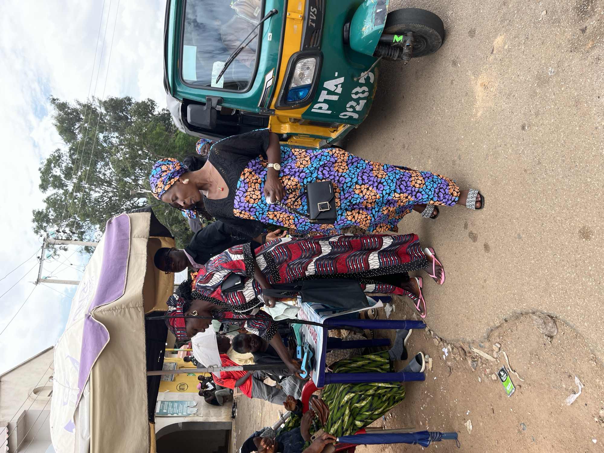 This image captures a bustling street scene in Jos, Nigeria, featuring a commercial transaction and passersby. In the foreground, a woman wearing a long, red, white, and black patterned dress with a matching headwrap stands, leaning over a small blue plastic table covered with a checkered cloth. She is holding a white cloth or paper in her left hand and reaching towards a blue electronic device on the table with her right hand, suggesting a transaction is in progress. She wears pink flip-flops and gold hoop earrings, with a black tote bag slung over her left shoulder. Seated to her left in a blue plastic chair is another woman dressed in a green and black patterned garment, engaged in the interaction at the table. A portion of a blue umbrella is visible next to her chair.
To the right of the central transaction, another woman walks past, facing left. She is adorned in a vibrant, multi-colored (purple, orange, blue, black) patterned long dress, a matching headwrap, dark slides with white patterns, a gold necklace with a red pendant, a gold watch, and a small black shoulder bag. Her mouth is slightly open, and she appears to be observing or speaking.
Behind the walking woman, a green and yellow TVS King three-wheeled auto-rickshaw (Keke Napep) with the registration "PTA-9285" is parked on an asphalt road. The street surface transitions from asphalt to a dirt shoulder in the foreground, where scattered debris, including plastic wrappers, is visible.
In the mid-ground, behind the transaction table, a large, light brown and purple market umbrella provides shade. Underneath and around it, several other individuals are present: a man in a black suit and sunglasses partially obscured, a person in a yellow shirt seated, and a child holding a small white umbrella. Further to the left, under what appears to be another canopy, additional people are visible, including one in a red shirt. The background features a light-colored building with an arched entrance, electrical poles and wires against an overcast sky, and a large tree with green foliage. The visible "MTN" logo on a sign in the background confirms a common telecommunications presence in Nigeria.