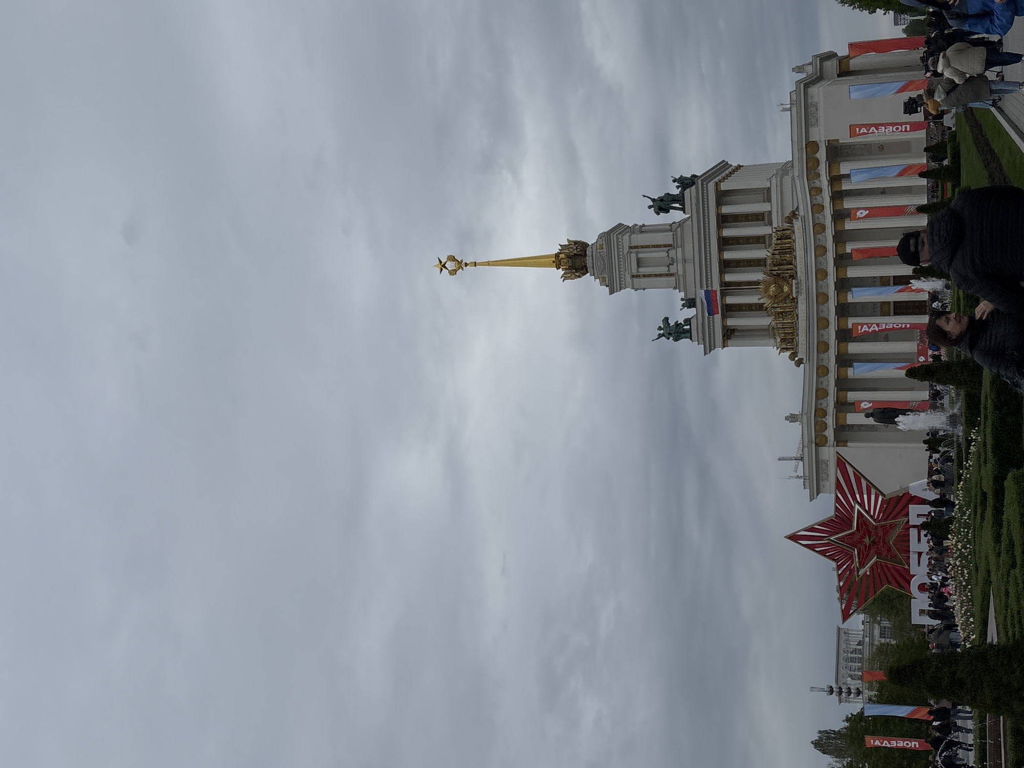 The image shows the Central Pavilion at VDNKh (Exhibition of Achievements of National Economy) in Moscow, Russia, on a cloudy day. The building is tall and white, with a golden spire topped with a star. There are sculptures on top of the building, as well as flags hanging down the side. A large red star is in the foreground. There are people walking around the area, including some near a fountain in front of the building. Vegetation is visible around the area, including bushes and trees. Banners with Cyrillic text are present.