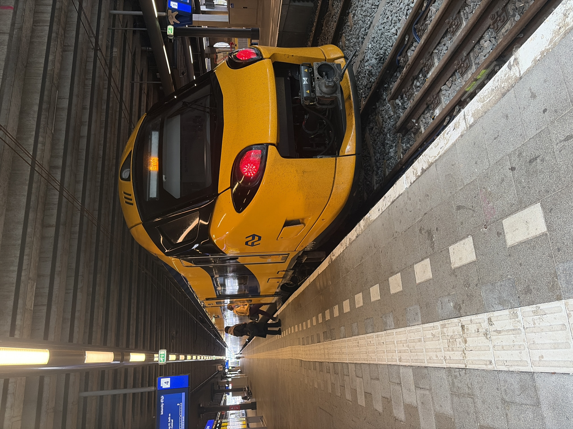 A bright yellow and blue train, identified as a Dutch Railways (NS) service by its livery and the double Z logo, is stopped at a station platform. The image is taken from the platform, looking towards the front of the train, which has its red tail lights illuminated. An access panel on the very front of the train is open, revealing internal components, suggesting maintenance or an operational check. A display on the front of the train shows partially visible text, which includes "Utrecht". The train number "3609" is visible on its side.
Two people are standing on the grey and white tiled platform next to the train. One person wears a yellow jacket, and the other is in dark clothing with a hat. The platform features a white tactile paving strip along its edge and square white markings further back.
The station environment appears to be either indoors or under a large cover, with a textured, concrete-like overhead structure and artificial strip lighting illuminating both the ceiling and the platform edge. In the background, blue information screens are visible, displaying "Intercity," the number "4" (likely a platform number), and destinations such as "Den Haag HS, Delft, Rotterdam Centraal." A green exit sign and another sign with the number "3" are also present. The train tracks, consisting of rails and ballast, are visible between the train and the platform. The scene captures a moment at a modern train station, possibly during a stop or a brief technical inspection.