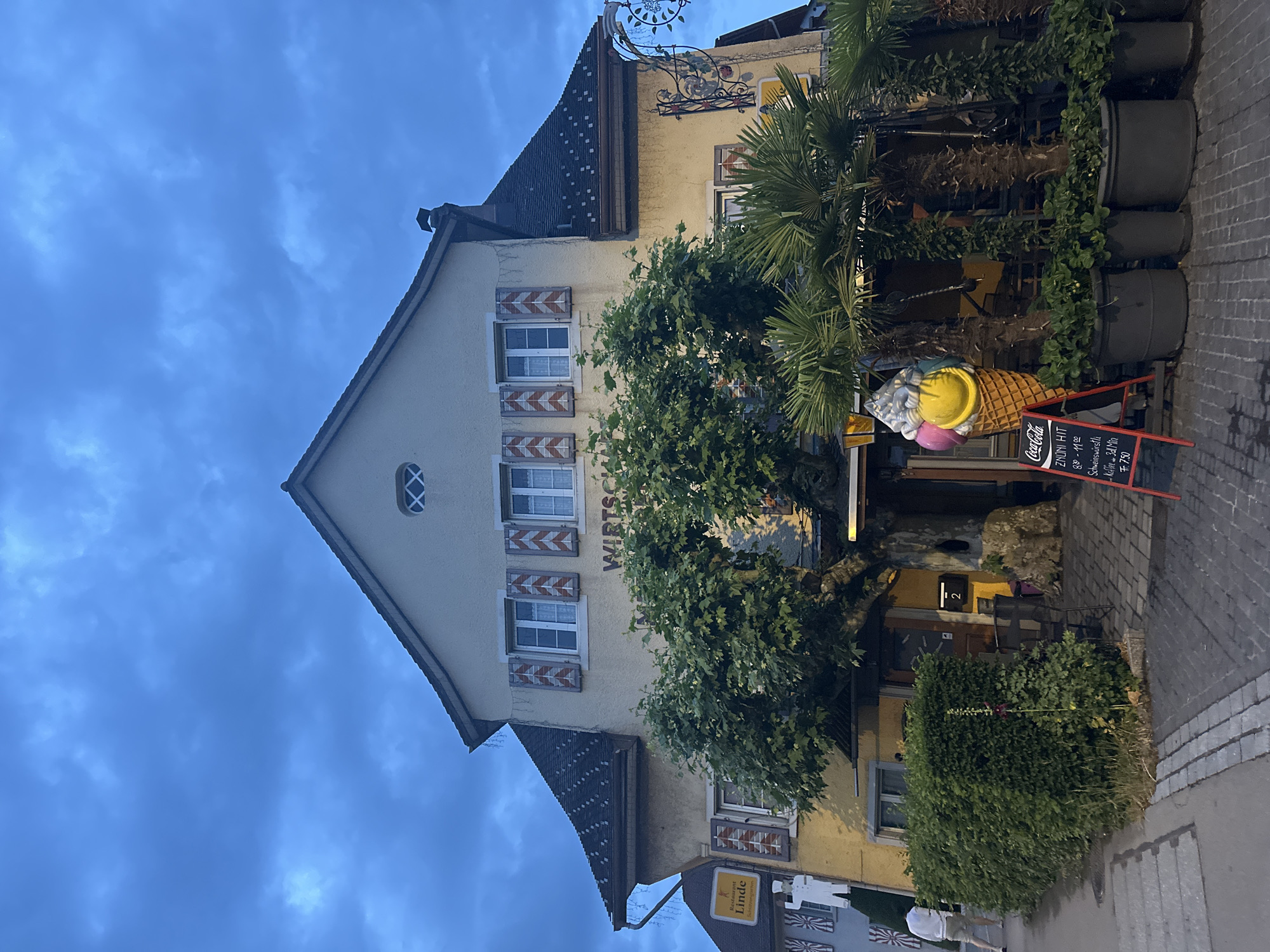 The image displays the facade of a multi-story building, likely a restaurant or inn, set against a deep blue, cloudy sky, indicating dusk or early evening in Embrach, Switzerland. The building has a light yellow stucco exterior, a dark tiled roof, and several windows adorned with distinctive red and white chevron-patterned shutters. A small, oval window with a cross pattern is visible on the gable.
Prominently featured are multiple signs and decorations. A decorative wrought-iron sign hangs from the upper part of the building. A yellow sign clearly reads "Restaurant Linde." A large, colorful ice cream cone sculpture, with yellow, pink, and white scoops, stands near the entrance, with "Coca-Cola" visible on its side. A red chalkboard sign on the sidewalk advertises "ZNÚNI HIT" (snack hit) from "8:00 - 11:00," listing "Schweinswürstli" (pork sausage) and "Kaffee" (coffee), priced at "Fr. 7.50." The word "WIRTSC" is partially visible on the building's facade. A house number "2" is also visible near an entrance.
Lush greenery, including a large tree with vibrant leaves, palm trees in pots, and ivy, adorns the front of the establishment, creating an inviting atmosphere. The sidewalk is paved with cobblestones. No people are visible in the scene.