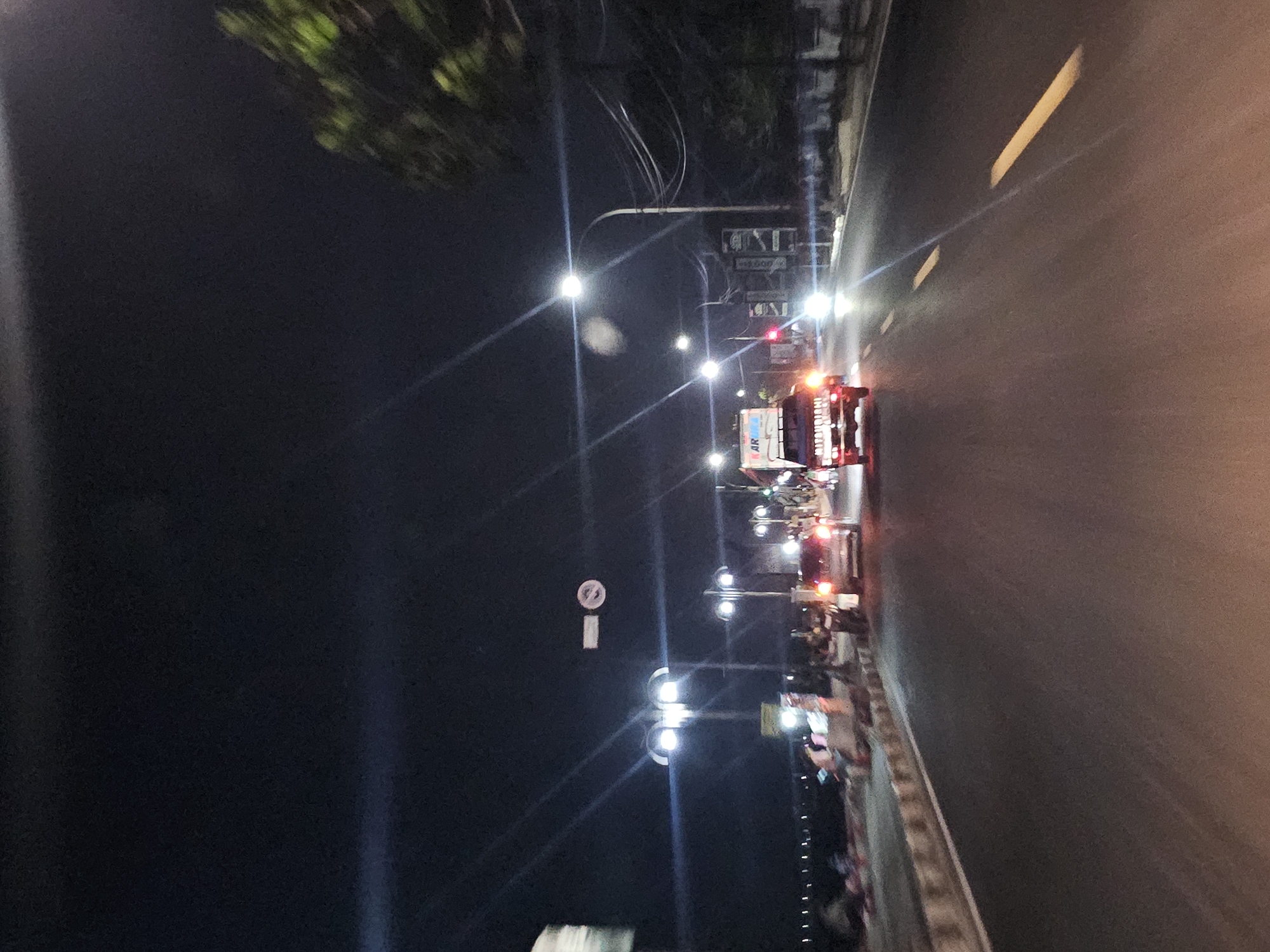 The image depicts a nighttime scene on a road in Tuban, Indonesia. The road is asphalt and marked with yellow lines, indicating lanes. Several vehicles are present, including a white truck with "AR" and "MITSUBISHI" visible on its side, and a silver car. Streetlights illuminate the scene, casting long streaks of light. On the left side of the road, there is a sidewalk with some pedestrians and motorcycles. Further left, a body of water is visible with lights reflecting on its surface. The sky is dark, suggesting it is night. Trees are visible on the right side of the road. Traffic signs are also present.