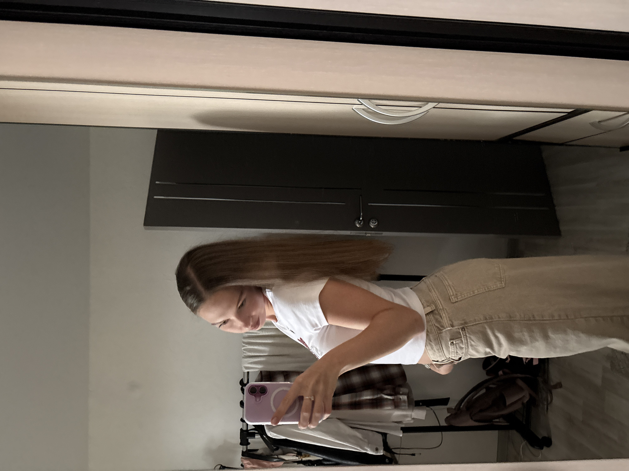 A young adult female with long, straight, brown hair, parted on the side, is depicted taking a mirror selfie in a residential interior located in Perm, Russia. She wears a white, short-sleeved crop top and high-waisted, wide-leg beige trousers. Her left hand, adorned with a ring on the ring finger, holds a light purple smartphone with a visible MagSafe ring, aimed towards her reflection in the mirror. She is slightly smiling and tilting her head.
To the left of the subject's reflection, a black metal clothing rack is visible, laden with various garments, including a white collared shirt, a brown and white plaid flannel shirt, and other dark clothing items. Behind the rack, white fabric or towels are draped over a bar. A brown backpack rests on the light-colored wooden floor beneath the rack. To the right of the subject's reflection, a dark brown door with a silver handle set is present, alongside a light beige wardrobe featuring two curved silver handles. The walls are light-colored, and the ceiling is plain.