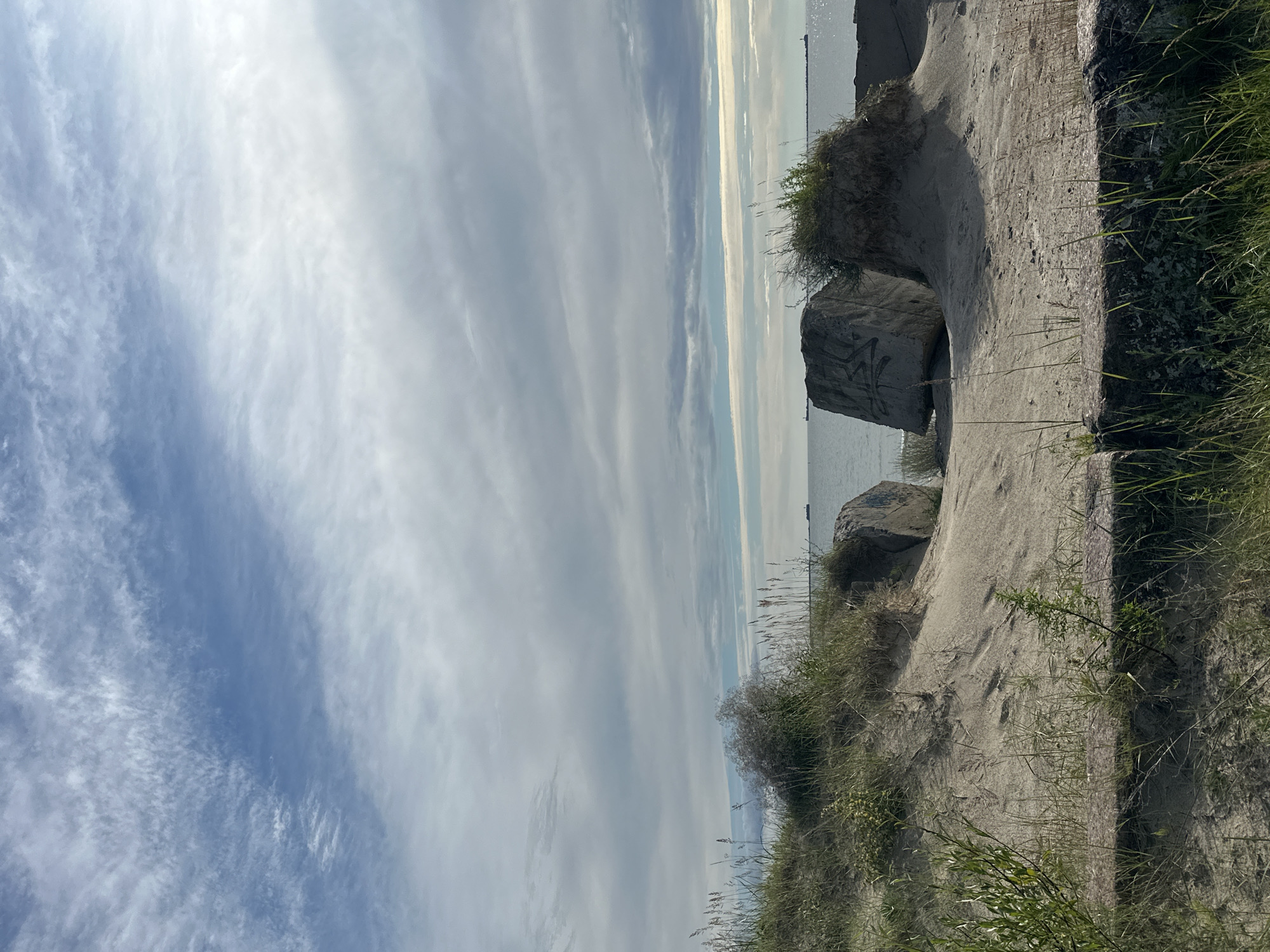 The image depicts a sandy embankment with sparse vegetation leading to the sea. In the foreground, there are rough, concrete-like structures partially covered in grass and sand, with some larger boulders visible. The sea stretches to the horizon, with a few distant ships. The sky is partly cloudy with a mix of blue sky and wispy white clouds. This scene is consistent with the beach area of Liepaja, Latvia, specifically near the breakwaters or coastal defense structures.