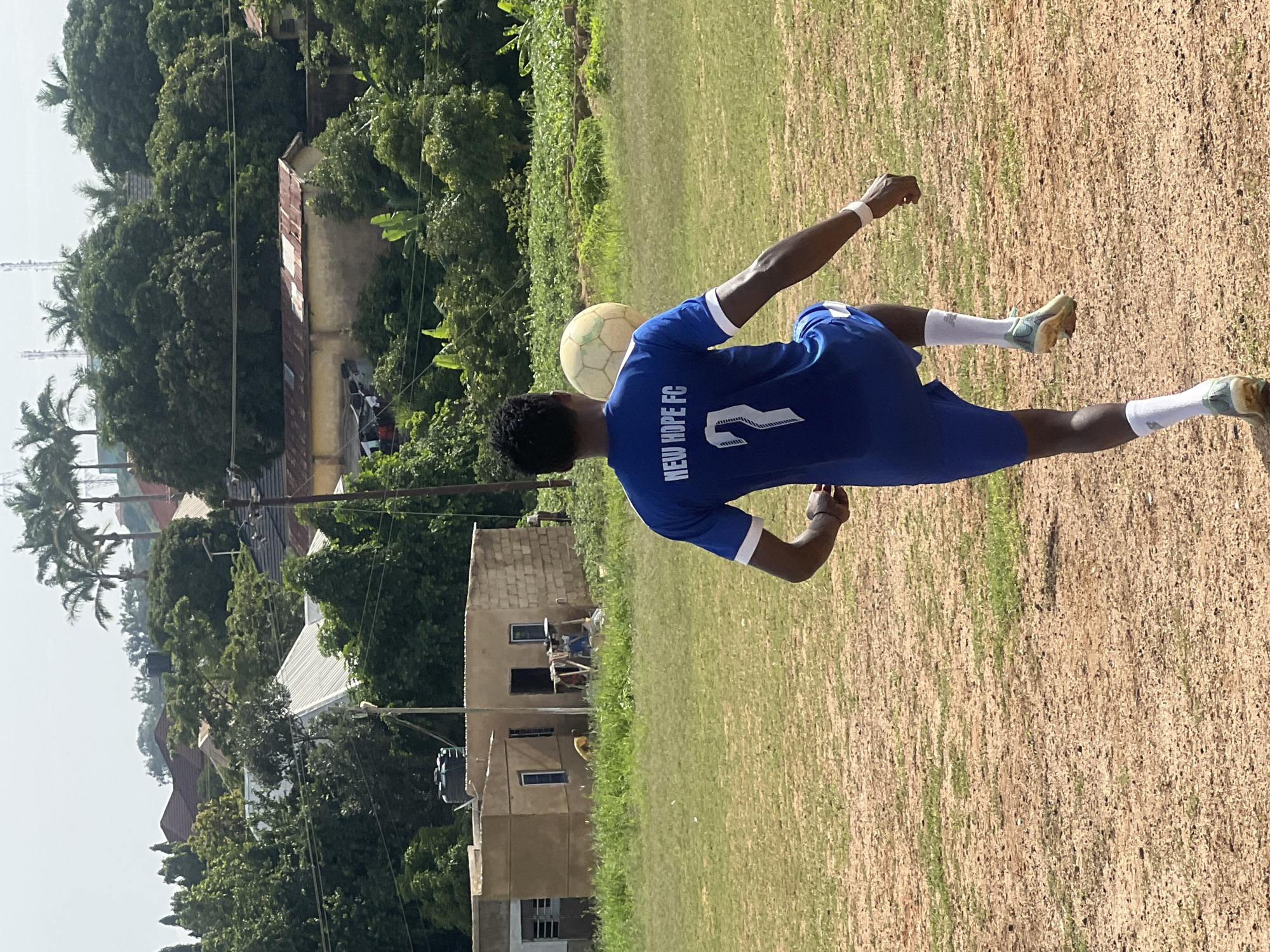 A young Black male wearing a blue "New Hope FC" soccer jersey with the number 7 and white socks is practicing with a soccer ball in a dirt field. He is in motion, with his right leg extended upward, balancing the ball on his thigh. The background shows a grassy field, trees, and some buildings, indicating an outdoor, rural or semi-rural setting. The environment appears to be warm and sunny, with a clear sky. The buildings in the background are simple structures, some with corrugated metal roofs. There are also palm trees visible, suggesting a tropical or subtropical climate. The image captures a moment of athletic training in a community setting.