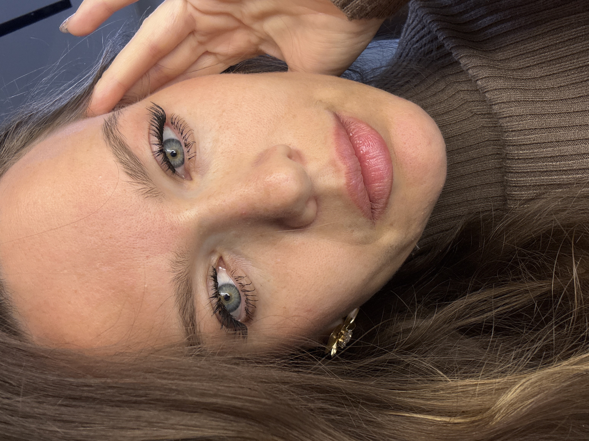 Here is a detailed description of the image:
The image features a close-up portrait of a woman with fair skin, light eyes, and light brown hair. She is wearing a brown turtleneck sweater. Her hair is styled with some highlights and is spread out around her head.
Her facial features are prominent: she has defined eyebrows, long eyelashes, and pink lips. She is wearing an earring. Her hand is placed near her forehead, with her fingers slightly touching her face.
The background is dark, possibly indicating an indoor setting. The lighting is focused on her face, highlighting her features. The image is shot at a slightly tilted angle, giving a unique perspective.