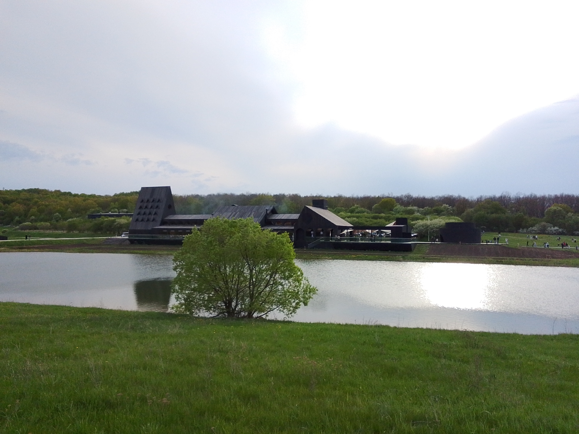 The image shows a body of water with a building and surrounding landscape in Алисово, Russia. The building is modern in design, with a black exterior and an angular, multi-level roof. There is smoke emerging from the building's chimney. The building is situated on the edge of the water, with a patio or deck area visible. The water reflects the sky and surrounding landscape. A small, green tree sits on the grassy bank in the foreground. The foreground also consists of a grassy area. In the background, trees cover the land. Some people are walking along the shore in the distance. The sky is overcast with bright clouds and sunshine breaking through.