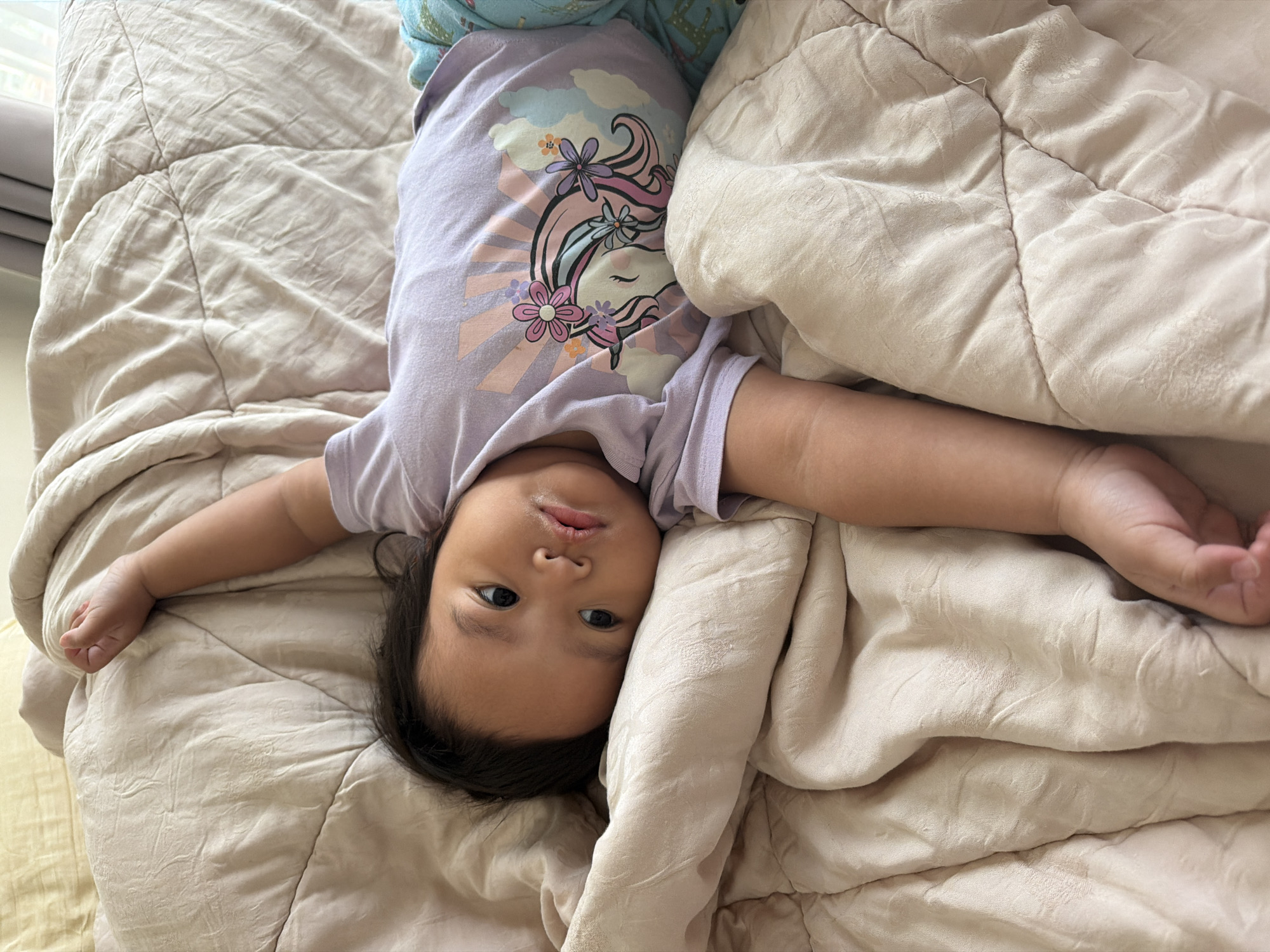 A young child with dark hair lies on their side on a light-colored, quilted blanket. The child is wearing a light purple t-shirt with a unicorn graphic and blue pajama bottoms. The child's right arm is stretched out above their head, while their left arm is bent and resting on the blanket. The child is looking towards the camera with a neutral expression. The background consists of more of the quilted blanket and a hint of a yellow pillow. There are no discernible elements to indicate a specific location in Central Jakarta, Indonesia.