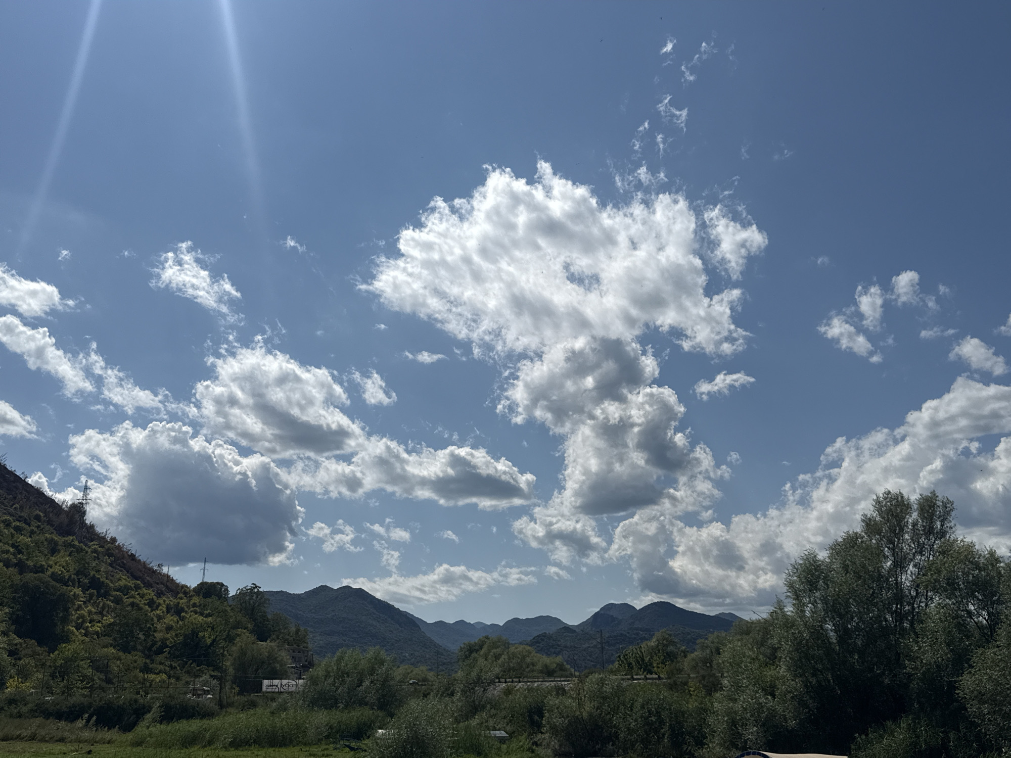 A wide-angle shot captures a bright, sunny day with a vibrant blue sky filled with fluffy white clouds. In the foreground, a lush green landscape stretches across the bottom of the frame, featuring various trees and foliage. The middle ground shows rolling hills covered in dense green vegetation, with several prominent mountain ranges receding into the distance. One of the hills on the left side of the image appears to have some areas of exposed earth or sparse vegetation, possibly indicating a slope. Power lines and towers are visible in the mid-ground, integrated into the landscape. The overall impression is a serene natural environment with hints of human infrastructure.