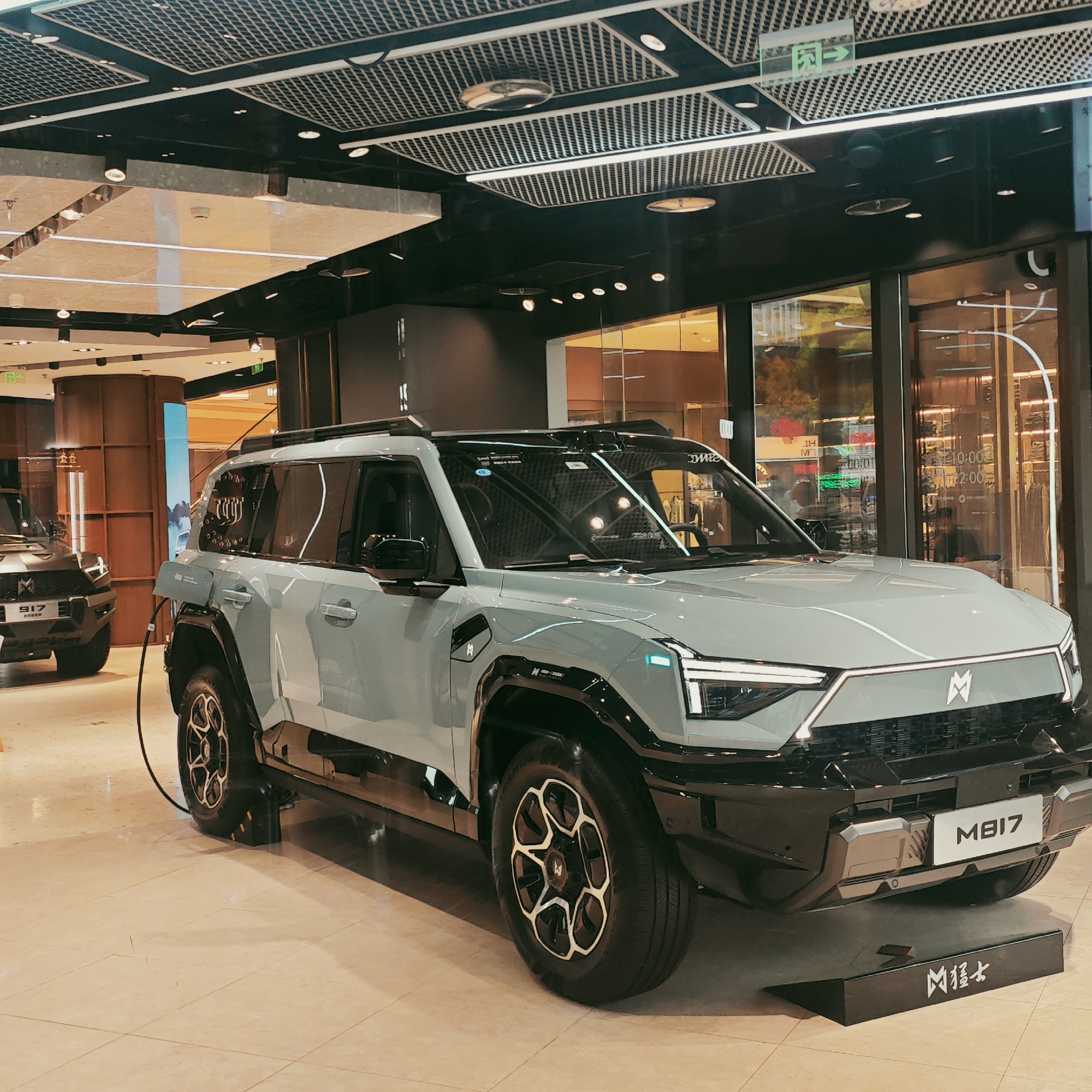 An indoor showroom or retail display in Yuzhong District, China, features two large, rugged-looking SUVs. The prominent vehicle in the foreground is a light grey (possibly matte finish) SUV, positioned at an angle, facing slightly left. It has a modern, angular design with distinctive headlights and a prominent "M" logo on its front grille. Below the grille, a plate displays "MB17". A black charging cable is connected to the vehicle's front left side. The SUV's large wheels have a unique geometric pattern. In front of this vehicle, a black display stand shows the Chinese characters "凶猛士" (Xiōngměngshì), which translates to "fierce warrior" or "ferocious gentleman," along with the "M" logo.
Partially visible in the left background is another SUV, dark grey or black, with a similar robust design and an "M" logo, displaying "917" on its front. The setting is brightly lit with various artificial lights, including spotlights and linear fixtures, reflecting off the polished light-colored tiled floor and large glass windows/walls in the background. The ceiling features a complex grid pattern with recessed lighting and green "exit" signs. Reflections of the interior and possibly outdoor elements are visible on the glass surfaces. Other visible text includes "IH" and "22:00" on signs in the background. The scene suggests a modern commercial space, likely a car dealership or a high-end mall, showcasing the vehicles.