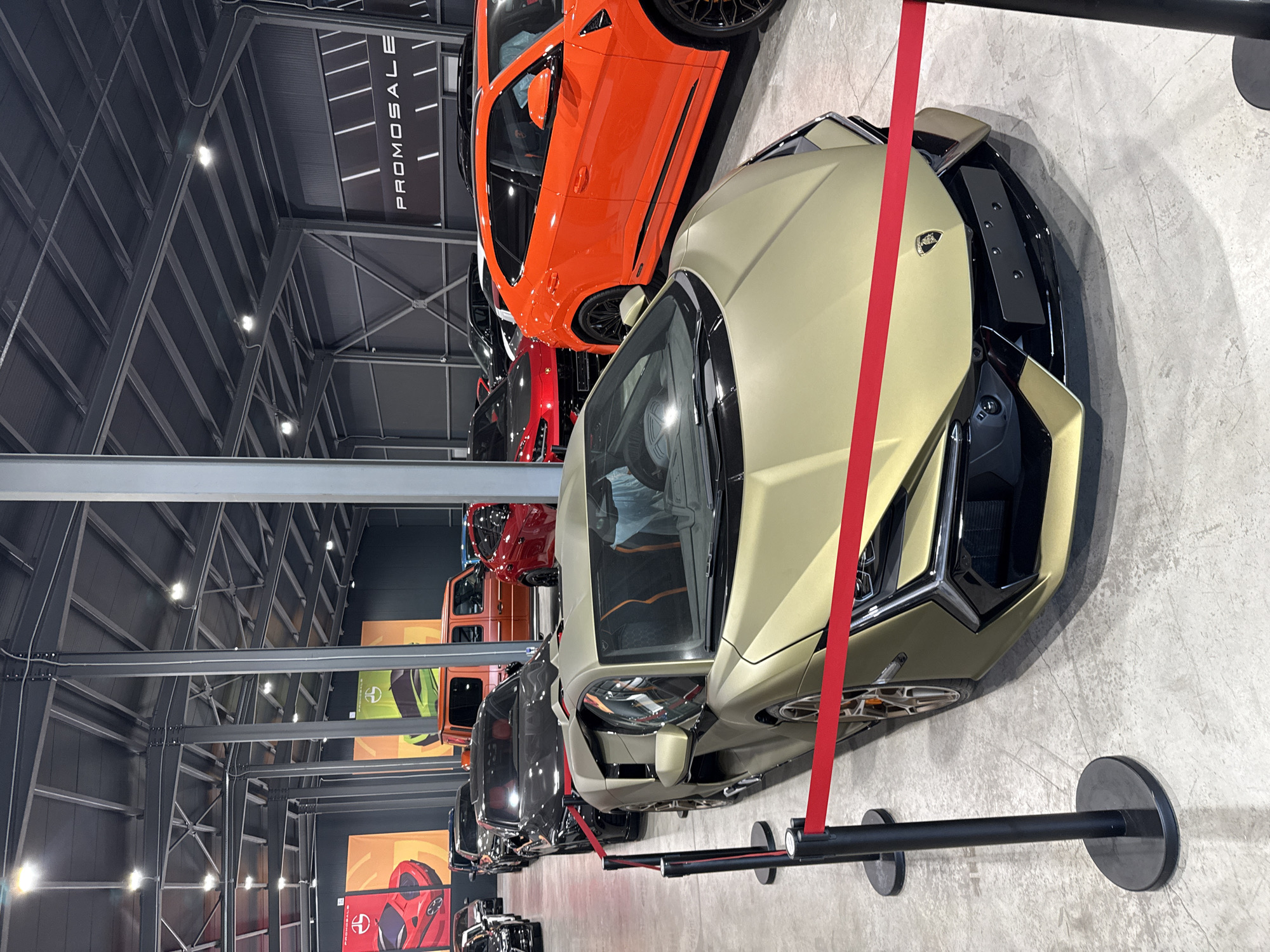 The image shows a showroom with several luxury cars, including a gold Lamborghini in the foreground. The showroom has a high ceiling with industrial lighting and exposed metal beams. Several black stanchions with red ropes are used to cordon off the cars. In the background, other cars are visible, including an orange Lamborghini Urus and a red sports car. Banners with car graphics and the text "PROMOSALE" are displayed on the walls. The floor is concrete. The scene is located in Plovdiv, Bulgaria. There are no people clearly visible in the image.