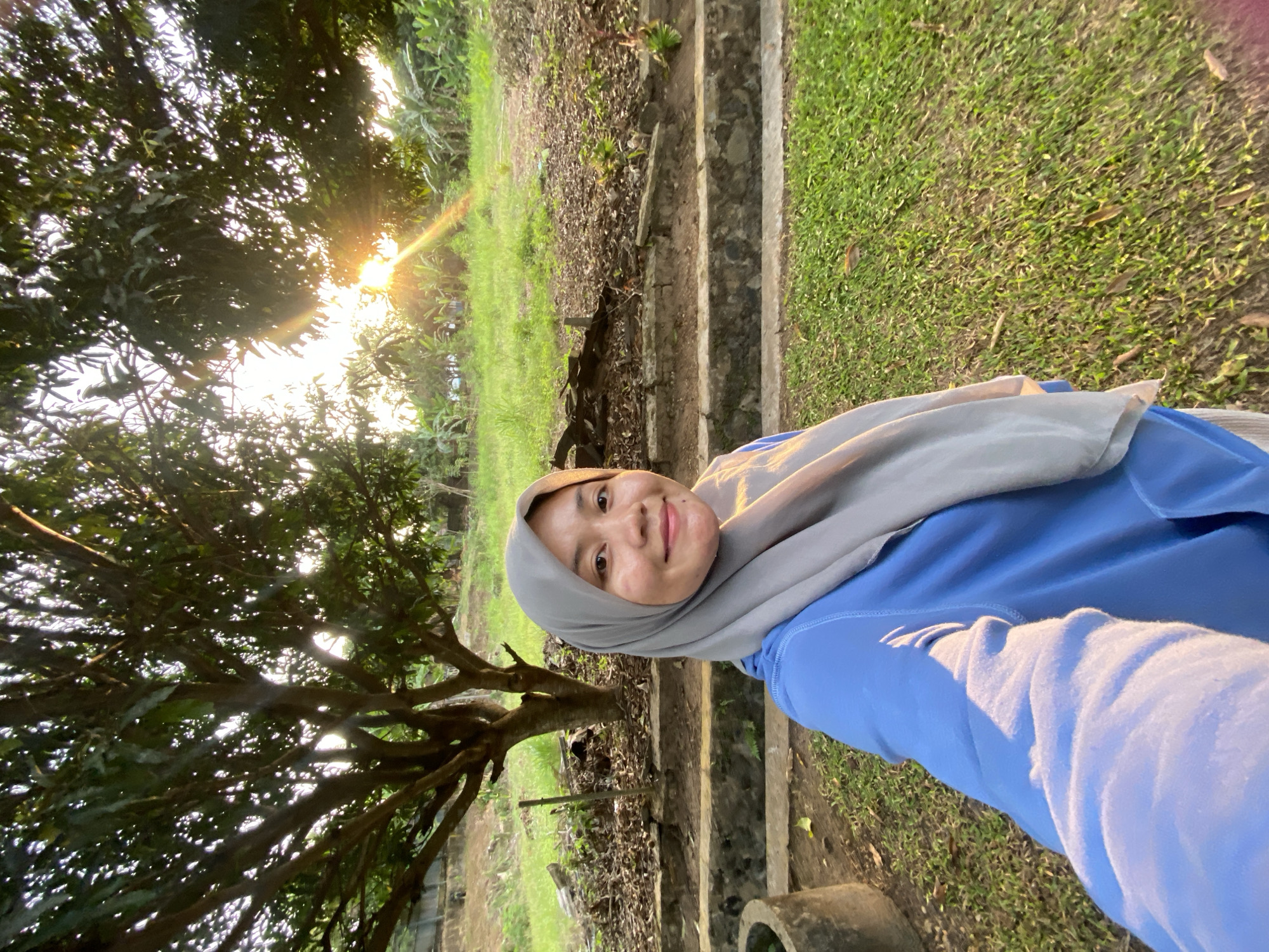 A woman wearing a light grey hijab and a blue long-sleeved shirt is taking a selfie in an outdoor setting. She is smiling and looking directly at the camera. Behind her, a large tree with dense foliage is visible, with sunlight filtering through the leaves and creating a lens flare. The background consists of a grassy area with some dry leaves scattered on the ground, a low stone wall, and further beyond, a field with banana plants and other vegetation. The sun is low in the sky, suggesting it is either sunrise or sunset, casting a warm glow on the scene. This location appears to be a natural or garden area, possibly in Banjarbaru, Indonesia, given the tropical vegetation.