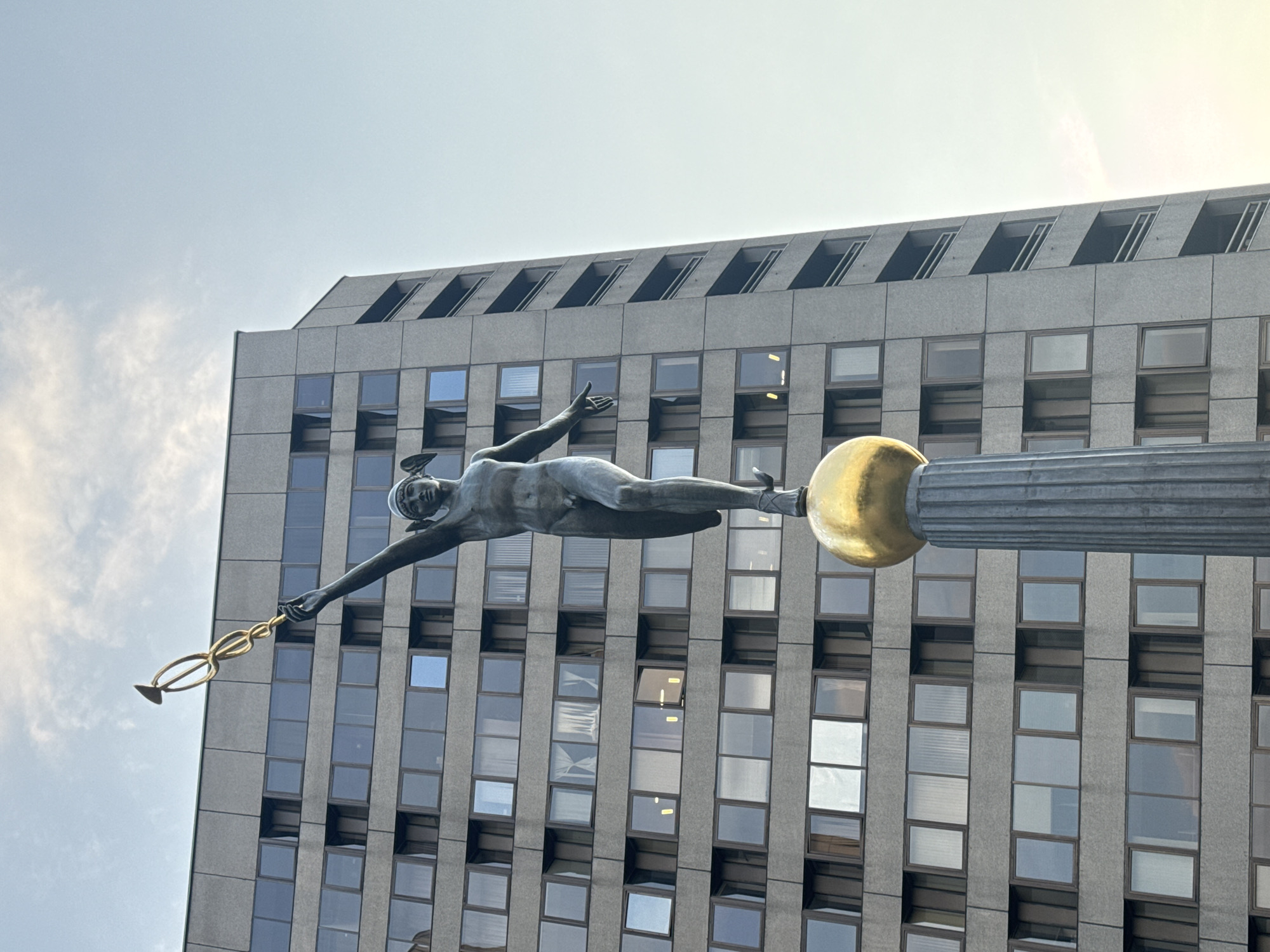 The image depicts the statue of Mercury at the top of the tower of the World Trade Center in Moscow, Russia. The statue is in flight, holding a caduceus. The background is a modern high-rise building with a grid of windows. The sky is visible in the upper part of the frame.