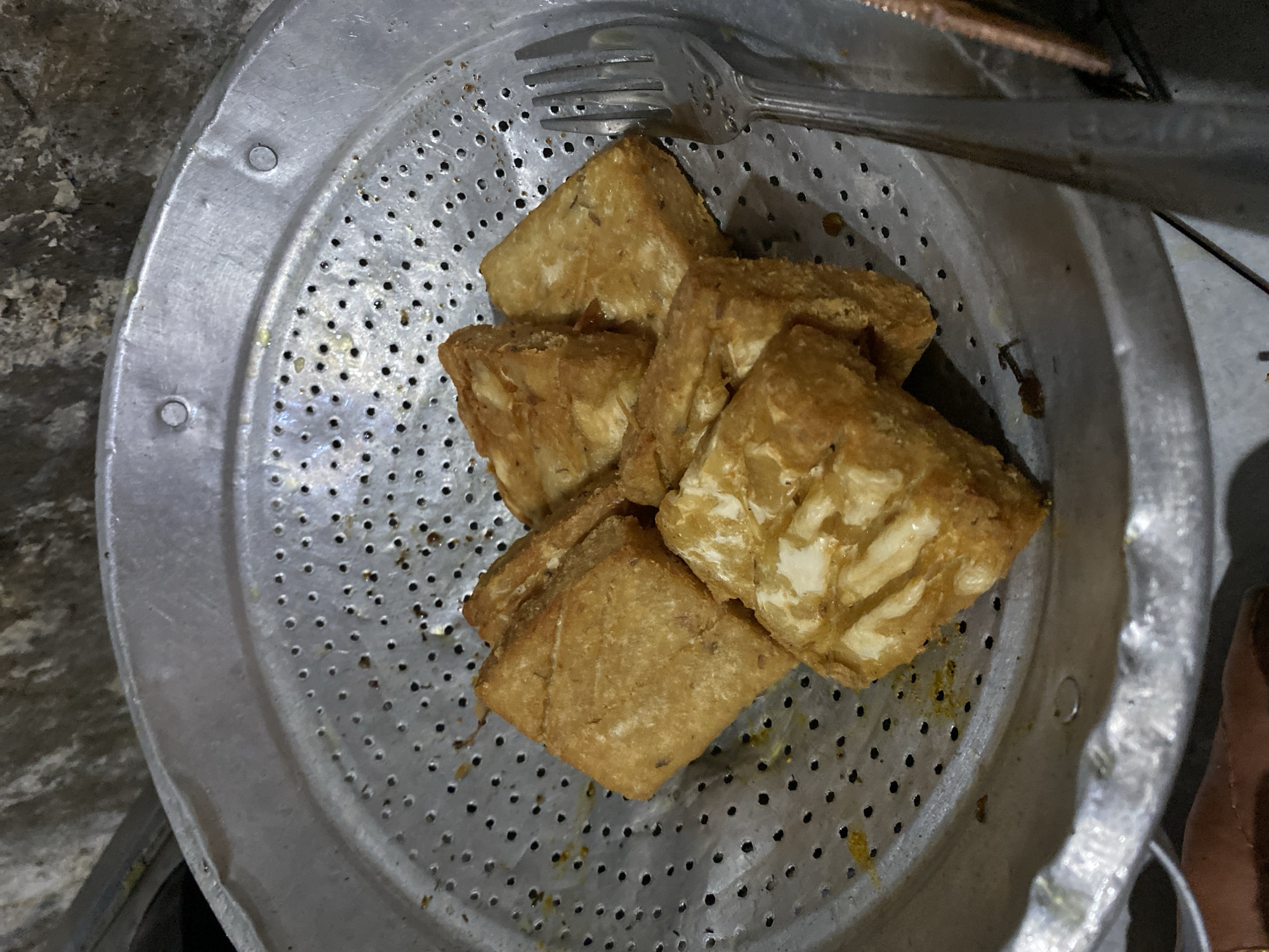 A close-up, overhead shot shows several pieces of fried tempeh on a perforated metal tray. The tempeh is golden brown and appears to be cut into rectangular pieces, with some pieces showing the layered texture of the fermented soybeans. A silver fork rests on the right side of the tray, its tines pointing towards the food. The tray itself is made of aluminum and has a patterned surface with many small holes. The background is out of focus, but appears to be a textured, possibly concrete or stone surface. The lighting suggests an indoor setting, possibly a kitchen or food stall, and the overall impression is one of simple, homemade food. Based on the popularity of tempeh in Indonesian cuisine, it is highly probable that this scene is located in Indonesia, potentially in the city of Madiun, known for its culinary traditions.