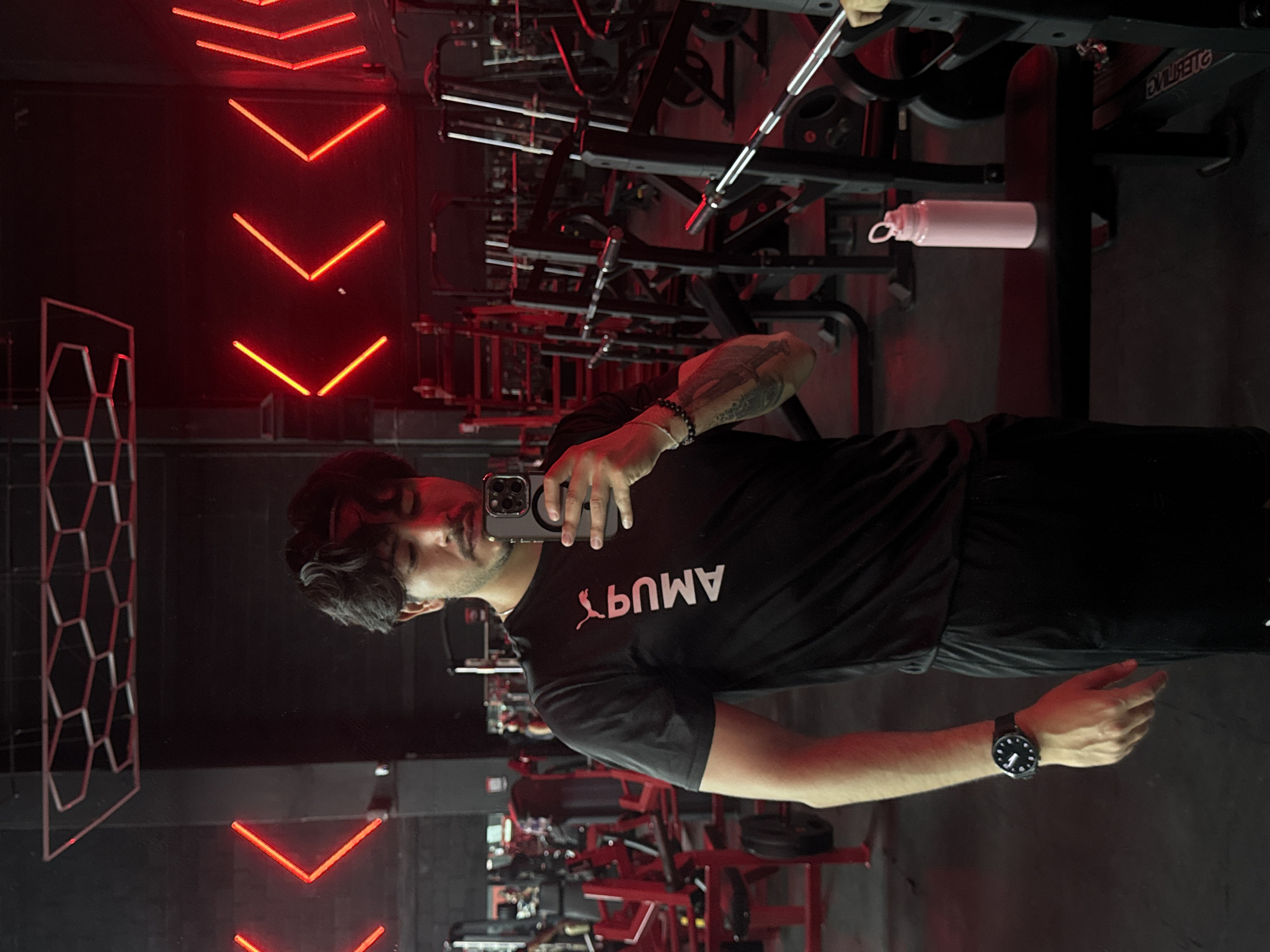 A person in a black t-shirt with white lettering is taking a selfie in a gym. The gym features red neon lighting, weightlifting equipment, and a pink water bottle. The photograph was taken in Puebla, Mexico. The person is wearing a watch on their left wrist and has a bracelet on their right wrist.