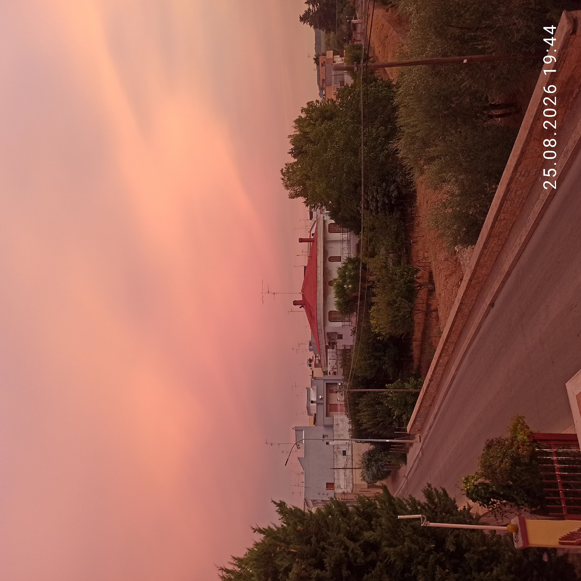 The image shows a street in Bitonto, Italy, with houses and trees lining the road. The sky is a soft pink, suggesting either sunrise or sunset. A red-tiled roof house is prominent in the center of the image. The date and time stamp at the bottom right corner reads "25.08.2026 19:44". There are no people visible in the frame. The scene depicts a quiet residential area, possibly at dusk.
