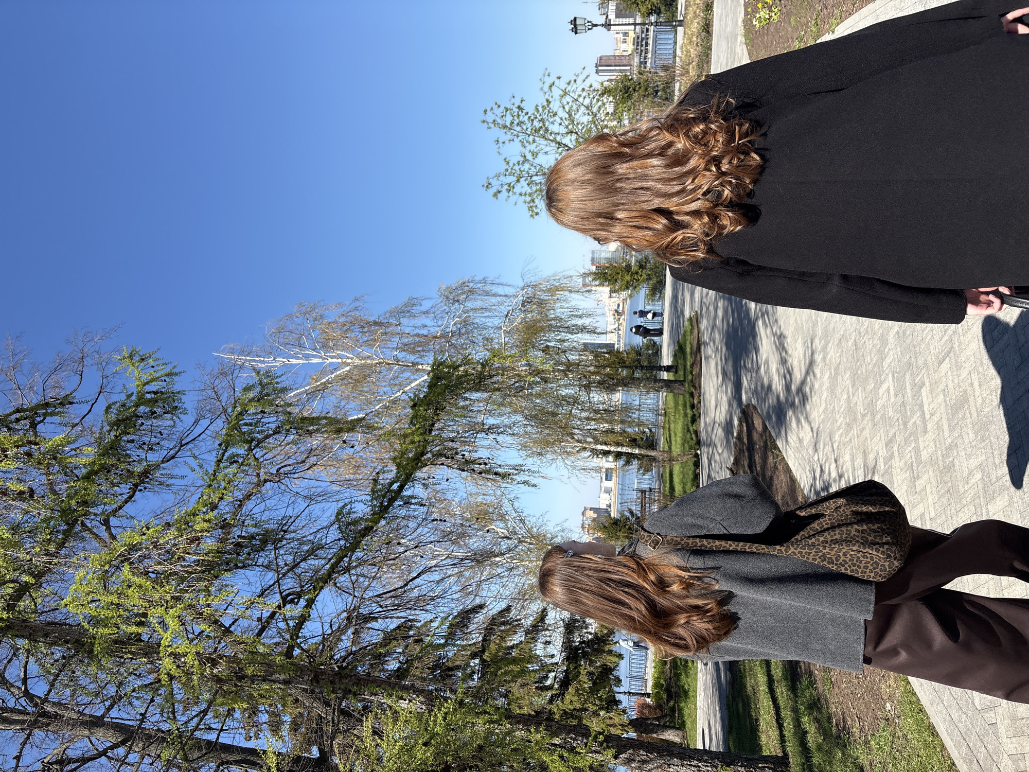 Two women stand on a paved path alongside a park in Kazan, Russia. The woman in the foreground wears a gray coat with a leopard-print bag and brown trousers. The woman behind her is wearing a black coat. Both have long, reddish-brown hair. The environment includes trees, a clear blue sky, and buildings in the distance near a body of water.