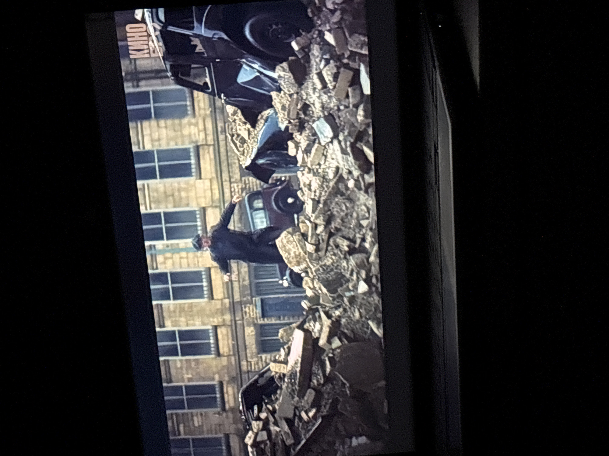 The image shows a scene from a screen, potentially at a cinema based on the word "KINO" visible in the upper portion of the screen. The scene depicts a car crash in front of a building with multiple windows. A man in dark clothing is visible, appearing to be moving amidst the debris and wreckage. The environment seems to be urban, with building elements and car parts scattered around. It is not possible to verify the location in the city of Budva, Montenegro with the details provided.