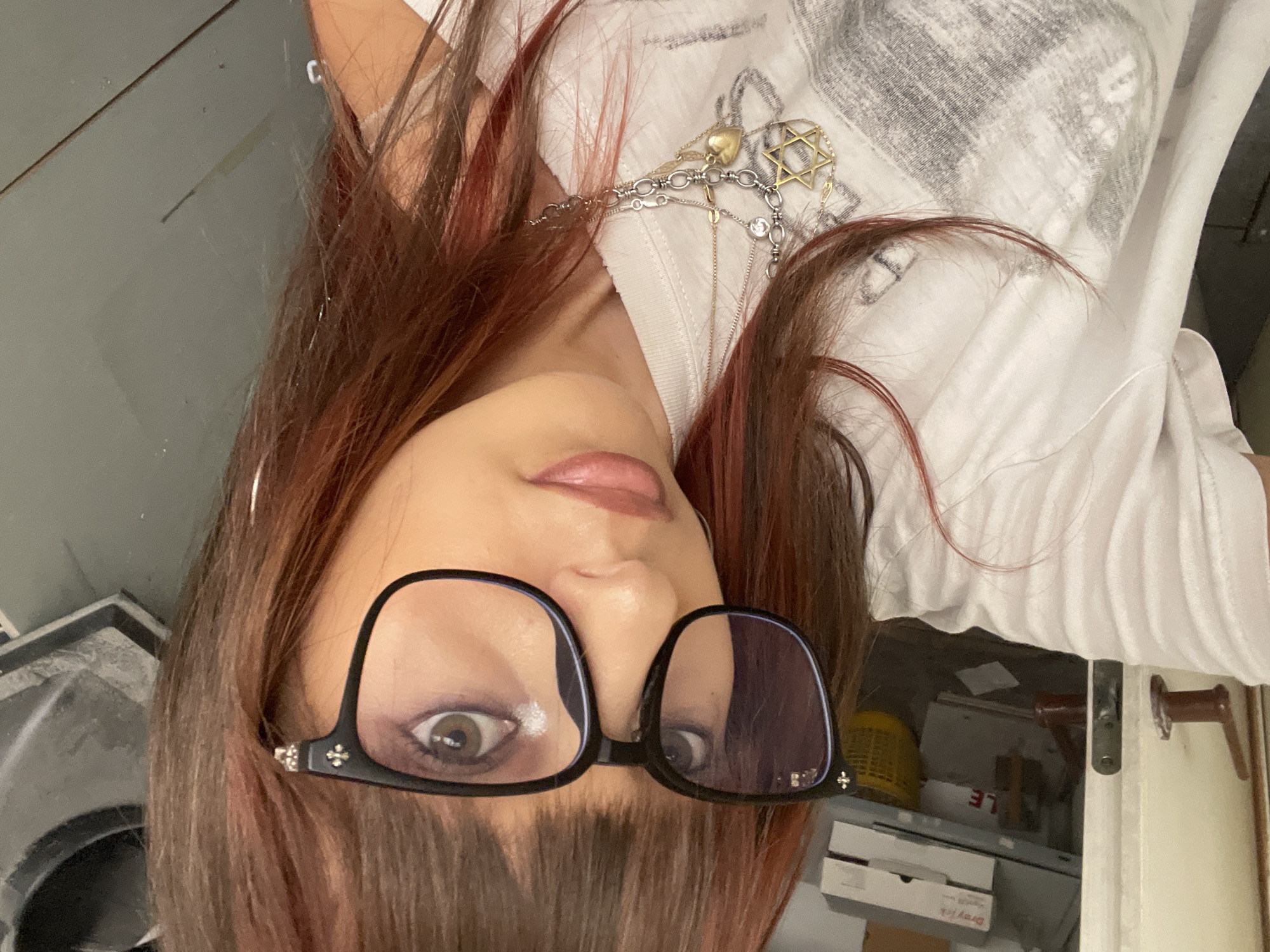 The image is a selfie taken by a young woman with reddish-brown hair, wearing black-rimmed glasses and a white t-shirt. She has on multiple necklaces, one featuring a Star of David pendant. Her makeup includes eyeshadow and lipstick. The background appears to be an indoor setting with some shelves and boxes, possibly in an apartment or storage area. The lighting is somewhat dim. There is no identifiable indication of the city of Raanana, Israel.