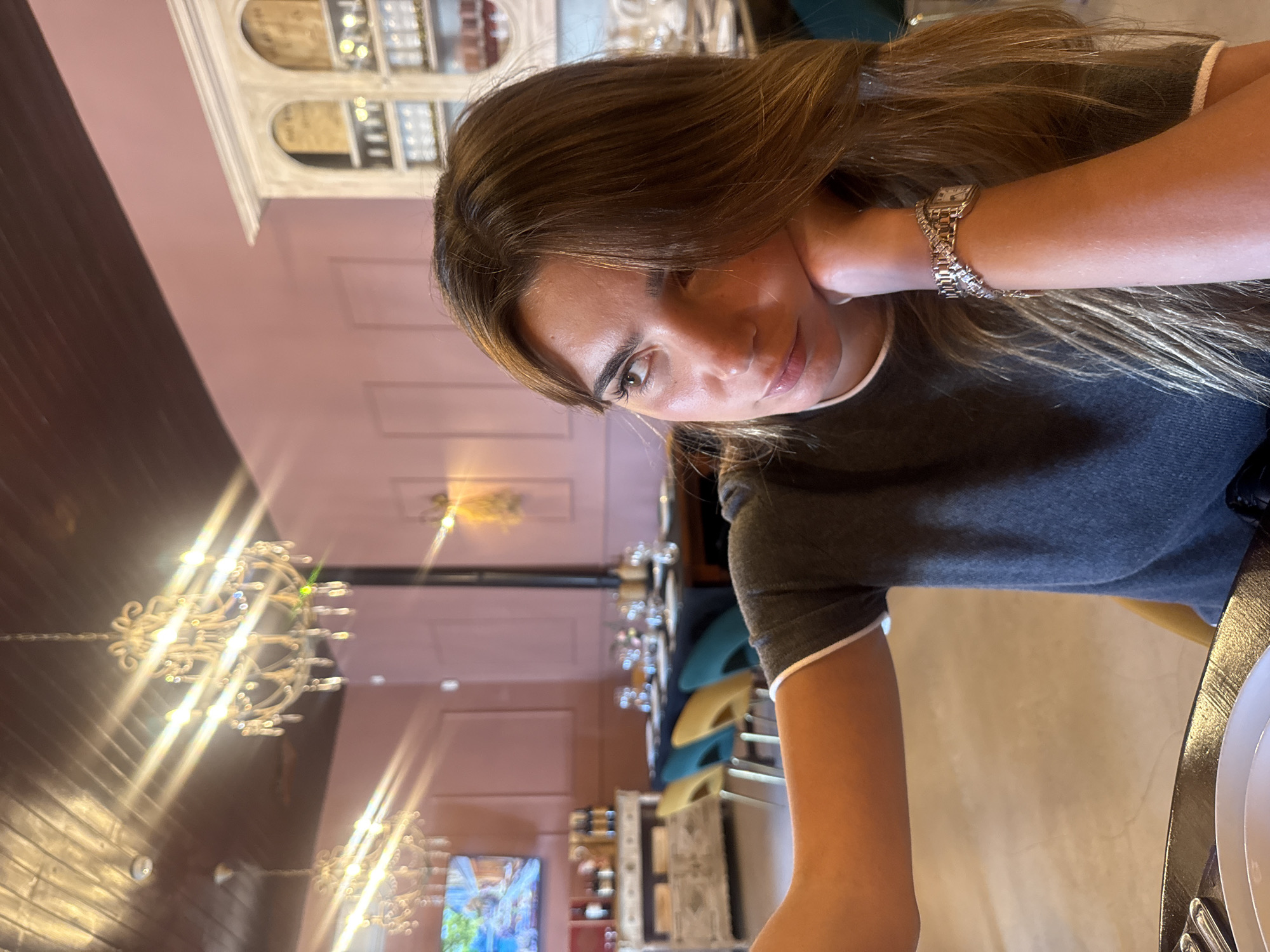 A medium shot, taken at a tilted angle, captures a young woman seated indoors, likely in a restaurant or cafe in Torres Vedras, Portugal. The woman, with long, straight brown hair, looks directly towards the viewer with a calm, slightly pensive expression, her head tilted to one side and her left hand resting gently near her face. She wears a dark grey t-shirt with white trim on the sleeves and collar. A silver-toned watch with a rectangular face and a matching, intricate metallic bracelet adorn her left wrist.
The background reveals a richly decorated interior. On the left, dark wooden paneling contrasts with pink-hued walls that extend into the mid-ground. Ornate, crystal-style chandeliers hang from the ceiling, emitting bright light flares that are prominent in the left and center of the frame. Decorative arched alcoves or shelving units, holding various items, are visible in the upper background. Further back, tables with white plates and cutlery can be seen, along with chairs in shades of turquoise and yellow. A television screen displaying colorful imagery is visible in the lower-left background. The overall lighting suggests an indoor setting, possibly evening, with reliance on artificial light sources. No discernible text is visible.