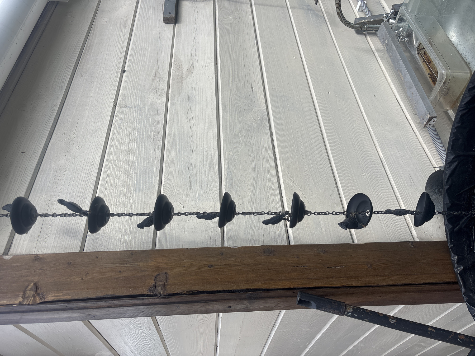 The image shows a close-up of a chain with decorative bird-shaped elements hanging against a white wooden wall. The chain is made of dark metal, with multiple cups and small bird figurines attached to it. To the left of the chain is a thick, weathered wooden post. The background is a horizontal planked wall, painted white. In the lower right corner, a sink or basin with some objects inside is visible, along with plumbing fixtures. The overall scene appears to be outdoors, possibly in a yard or on a patio, given the wooden structures and outdoor elements. The specific location of Лотос Снт (марьино), Russia, is provided as context but not visually depicted in the image itself.