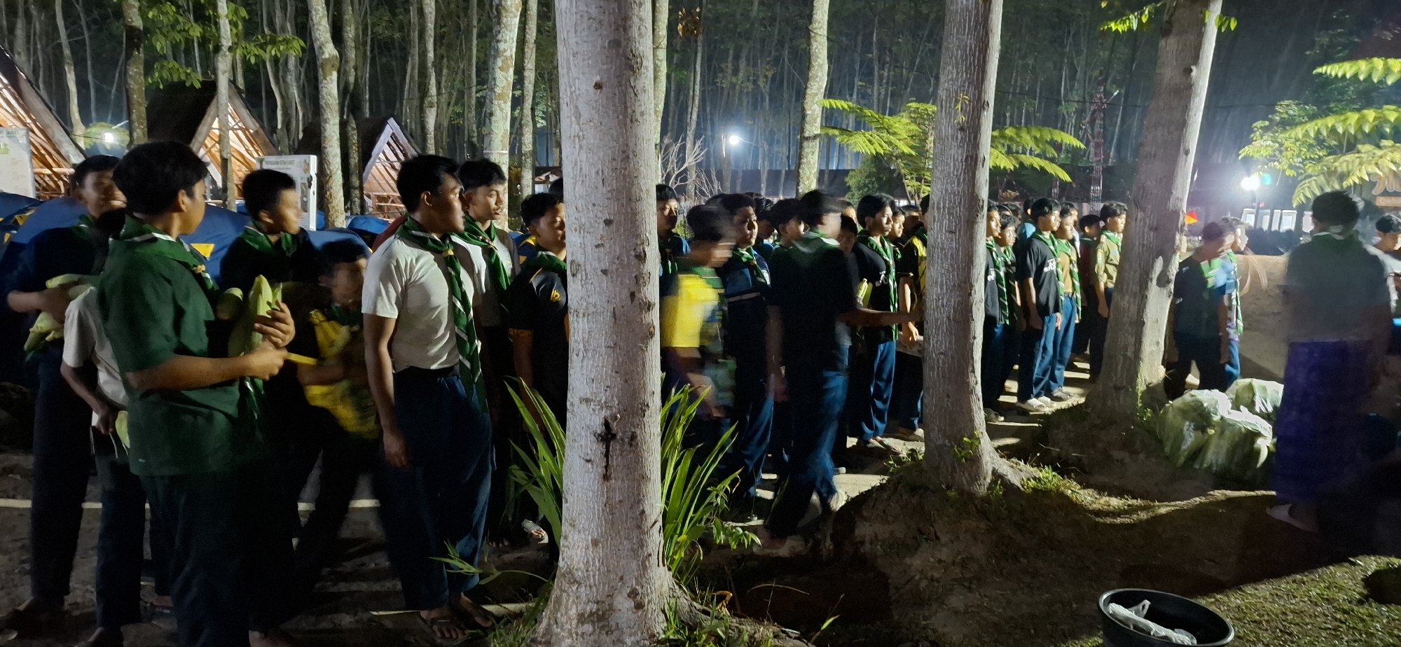 In Puntukrejo, Indonesia, at night, a group of young people, likely scouts due to their matching green scarves, stand in rows amongst trees. Tents are visible in the background, illuminated by artificial lights. The individuals are wearing dark shirts and pants, with some holding what appear to be green food items. The scene suggests an outdoor camp or gathering.