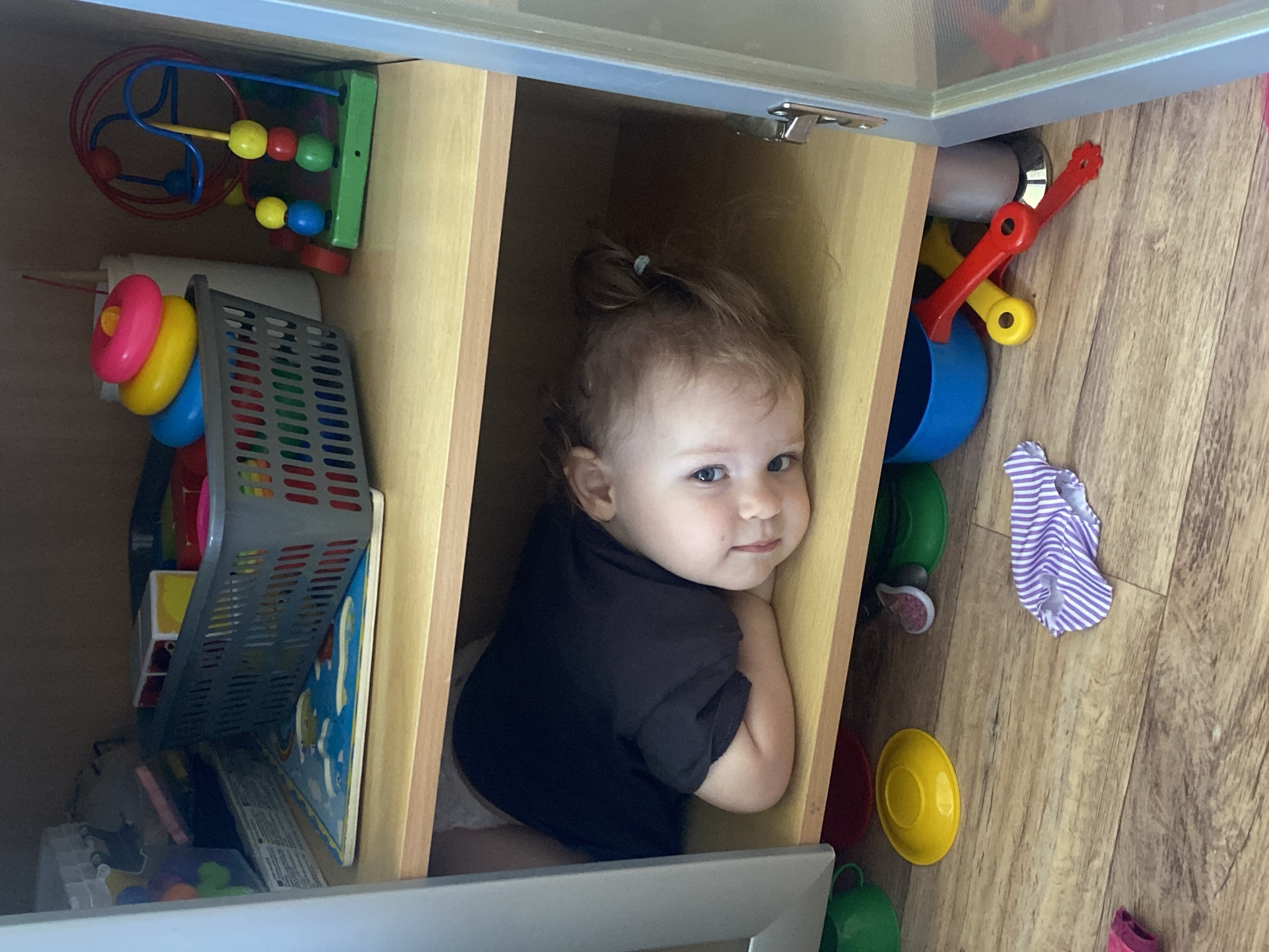 An overhead shot captures a playful baby nestled inside a light wooden cabinet compartment, surrounded by toys. The baby, with light brown hair tied in a small top bun and striking blue eyes, gazes directly at the camera with a sweet, knowing smile, embodying a moment of innocent mischief. They are wearing a simple black t-shirt.
The cabinet is sectioned into multiple compartments, all filled with children's playthings. To the baby's immediate left, within the cabinet, are a colorful bead maze featuring red, yellow, and blue beads on a wire, and a green base. Below this, a stacking ring toy with pink, yellow, and blue rings is visible next to a gray plastic basket that holds more items, including what appears to be a puzzle or book.
Outside the cabinet, to the baby's right, a collection of toys spills onto a light-colored wooden floor. These include a bright blue cup, red and yellow plastic scoops, green and yellow plates or bowls, and a small, striped purple and white piece of fabric, possibly doll clothing. The scene is brightly lit, suggesting it's daytime, and the overall atmosphere is one of a cozy, toy-filled play area in a home. No text is visible in the image.