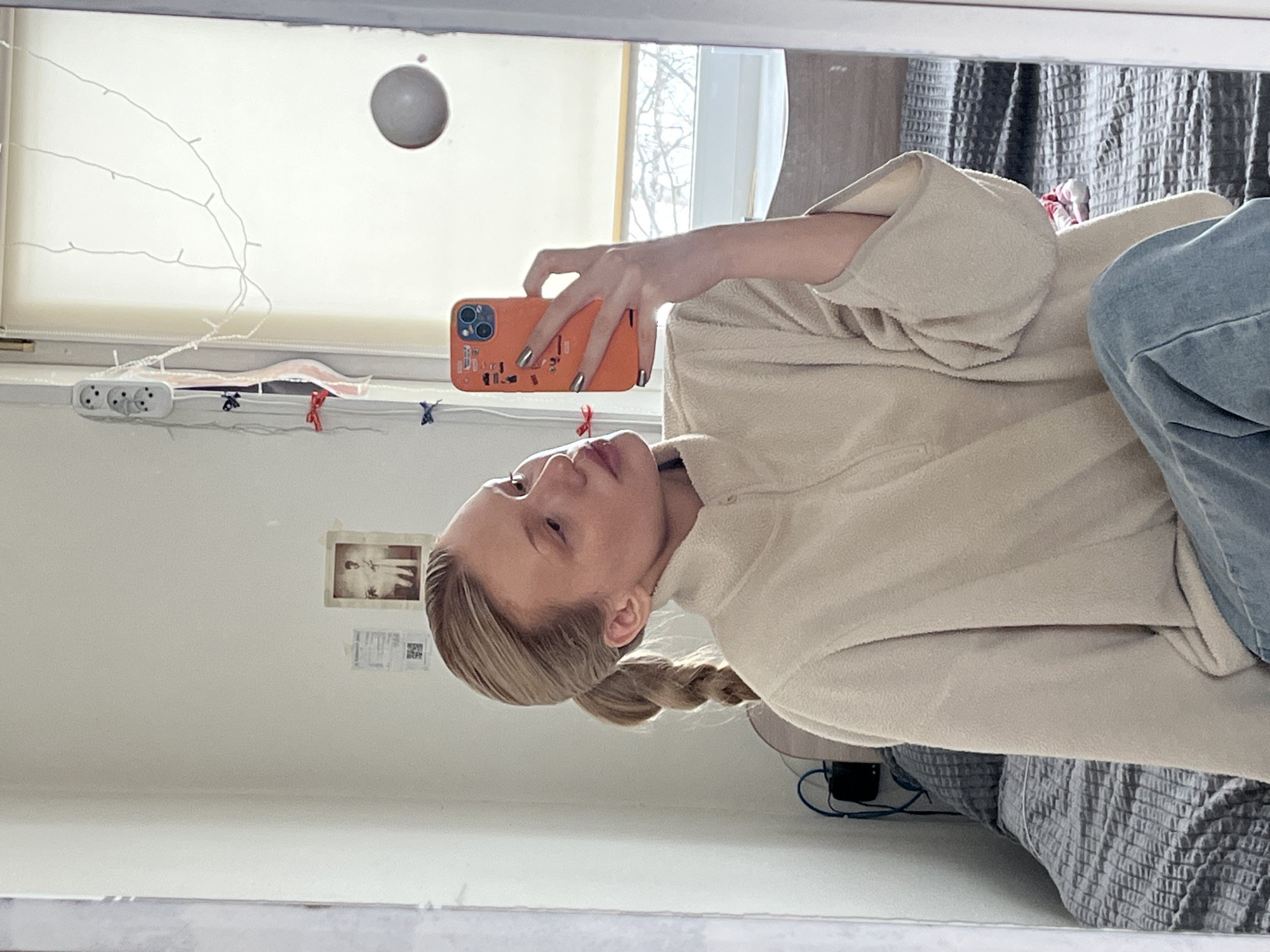 A young woman with blonde hair styled in a braid is taking a selfie in front of a mirror. She is holding an orange mobile phone. She is wearing a beige, fleece-like top and blue jeans. The background includes a window with a partially visible cityscape, possibly in a cold climate with leafless trees. A white roller blind is covering the window. An electrical outlet with multiple plugs and a string of decorative lights are positioned on the window sill. A small framed picture is mounted on the wall, along with taped notes. The setting appears to be an apartment interior. It is not possible to determine if the image was taken in Chaikovsky, Russia from the given information.