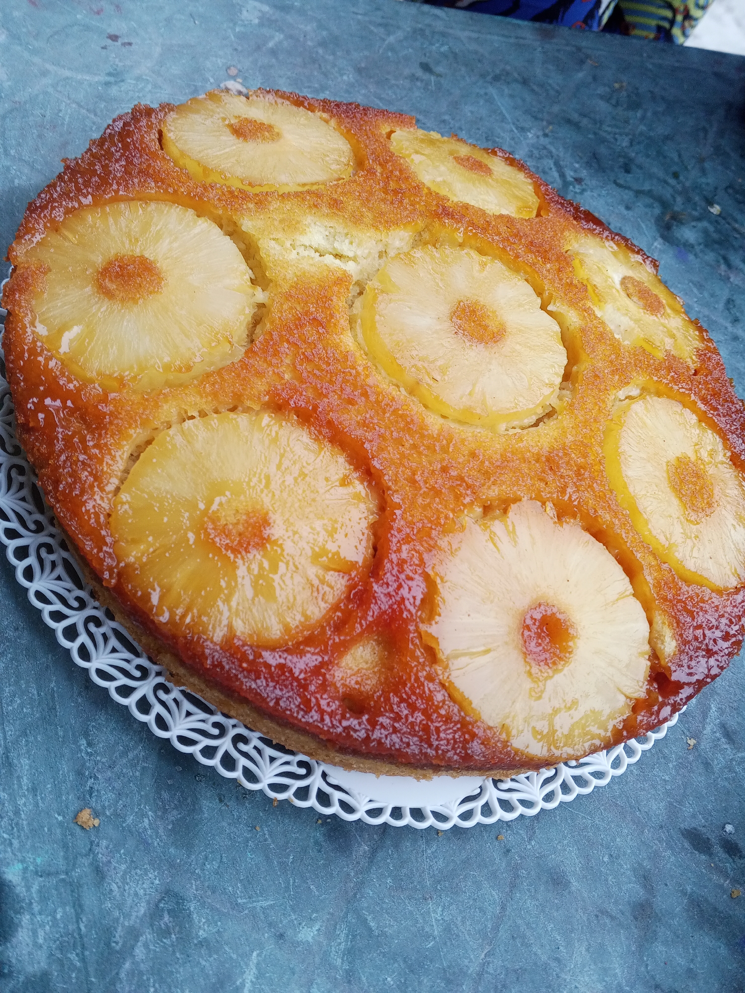 The image shows a pineapple upside-down cake on a white, decorative paper doily. The cake consists of a golden-brown sponge base with caramelized pineapple slices arranged in a circular pattern on top. The cake is positioned on a gray, mottled surface. The location is in the city of Tinaquillo, Venezuela. The image appears to be taken in an indoor setting with natural lighting.