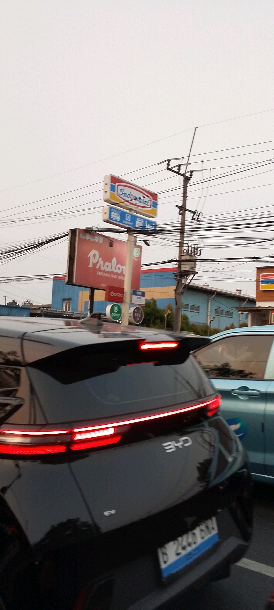 The image captures a street scene in Depok, Indonesia, likely during daylight hours, given the bright sky. The foreground features the rear of a black electric vehicle, possibly a BYD, indicated by the "BYD" emblem on its trunk and an "EV" badge. The car's taillights are illuminated, showing a red light bar across the rear and red brake lights. A blue license plate, partially visible, reads "B 2440 BXA". To the right, a light blue car is partially visible, suggesting a traffic or parking situation.
In the background, a utility pole with numerous electrical wires is prominently displayed. Several signs are visible, including an "Indomaret" convenience store sign with the brand's distinctive logo, and a large red sign with white text that reads "Pralon PERTAMA DAN TERBAIK". Below this, smaller signs include "OCBC" and ATM indicators. Further in the background, blue and yellow buildings are visible, some appearing to be commercial establishments. The overall environment suggests an urban or suburban setting with active commercial activity and infrastructure.