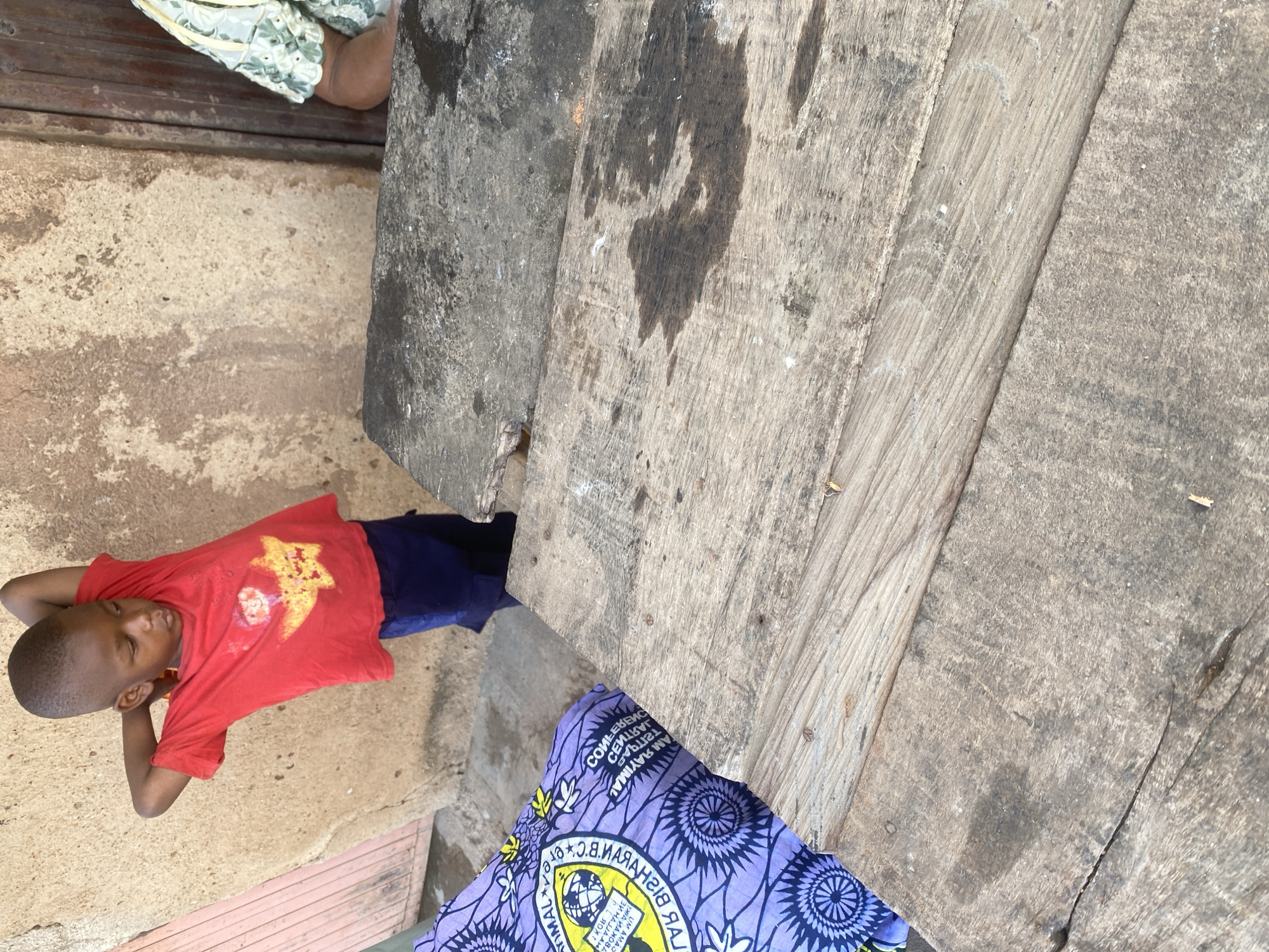 A young boy in a red t-shirt and blue shorts lies on the ground, resting his head on his hands. He appears relaxed, with his eyes closed. The t-shirt has a yellow star design on it. Beside him, a piece of purple fabric with a circular pattern is visible. The scene is outdoors, with weathered wooden planks and a rough concrete surface forming the ground and an elevated structure. The lighting suggests it is daytime. The overall setting appears to be a casual, possibly rural or semi-urban environment in Jos, Nigeria. There is some text visible on the purple fabric, but it is not clearly legible.