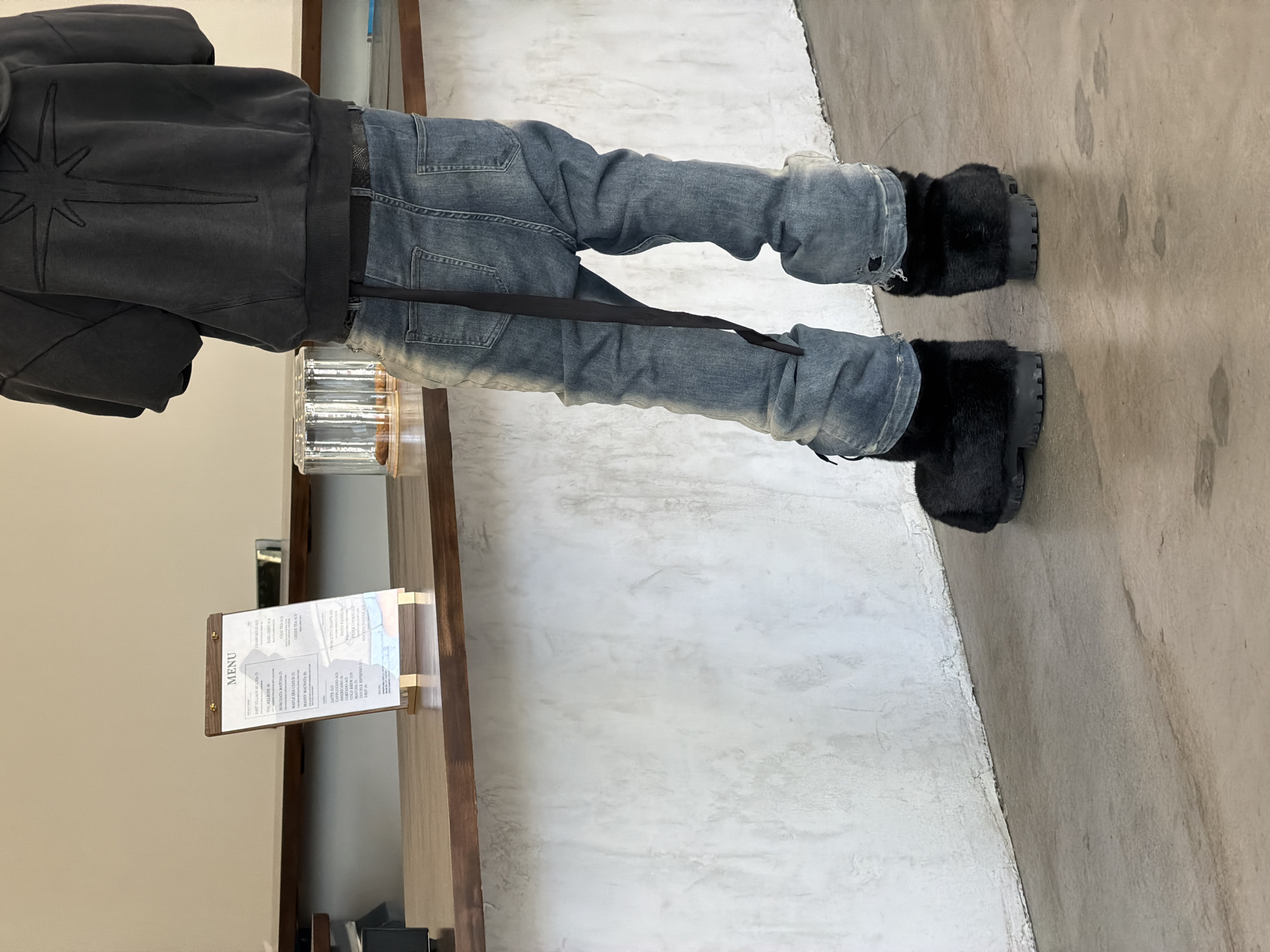 A person stands next to a dark-stained wooden counter with a menu. The menu is printed on a white paper in a wooden frame. The person wears a dark grey jacket with a large star emblem on the back, faded blue jeans with a suspender, and large black fur-covered boots. The boots are on a concrete floor. The scene is likely indoors, near a counter where a customer can order from a menu. The location cannot be definitively determined based on the provided image.